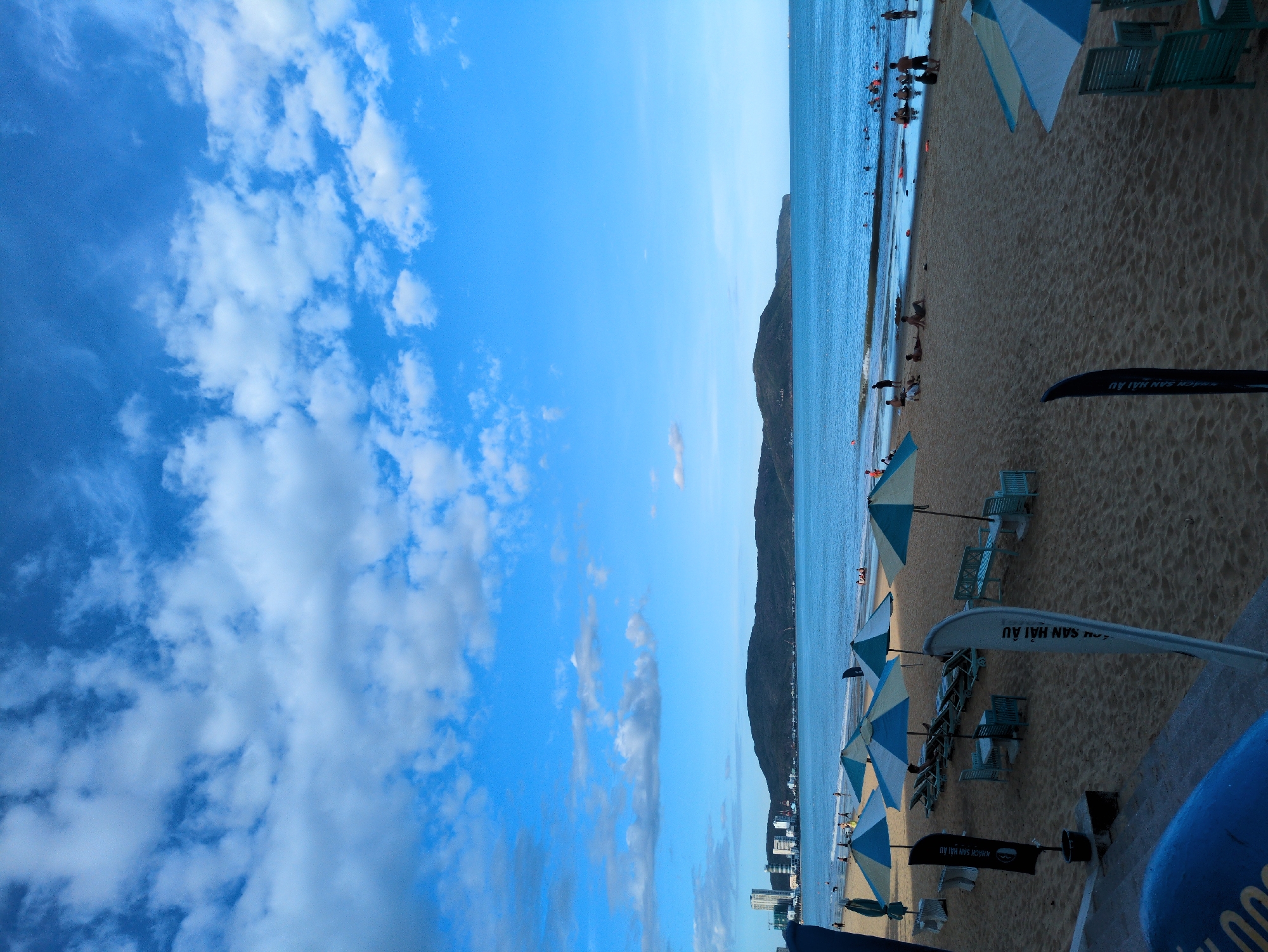 This image depicts a wide, sandy beach scene on a sunny day with a bright blue sky filled with scattered white clouds. In the foreground, several beach umbrellas, some open and some closed, are set up on the sand, along with lounge chairs and benches. Two tall, black and white flags with text in Vietnamese stand prominently on the sand. The middle ground shows the calm, blue ocean with several people swimming and relaxing in the water. In the background, a city skyline with tall buildings is visible along the coastline, bordered by a dark green, mountainous landscape. The overall impression is a vibrant and active beach environment. This scene is likely located on the main beach of Quy Nhon, Vietnam, which is known for its beautiful coastline and city views.
