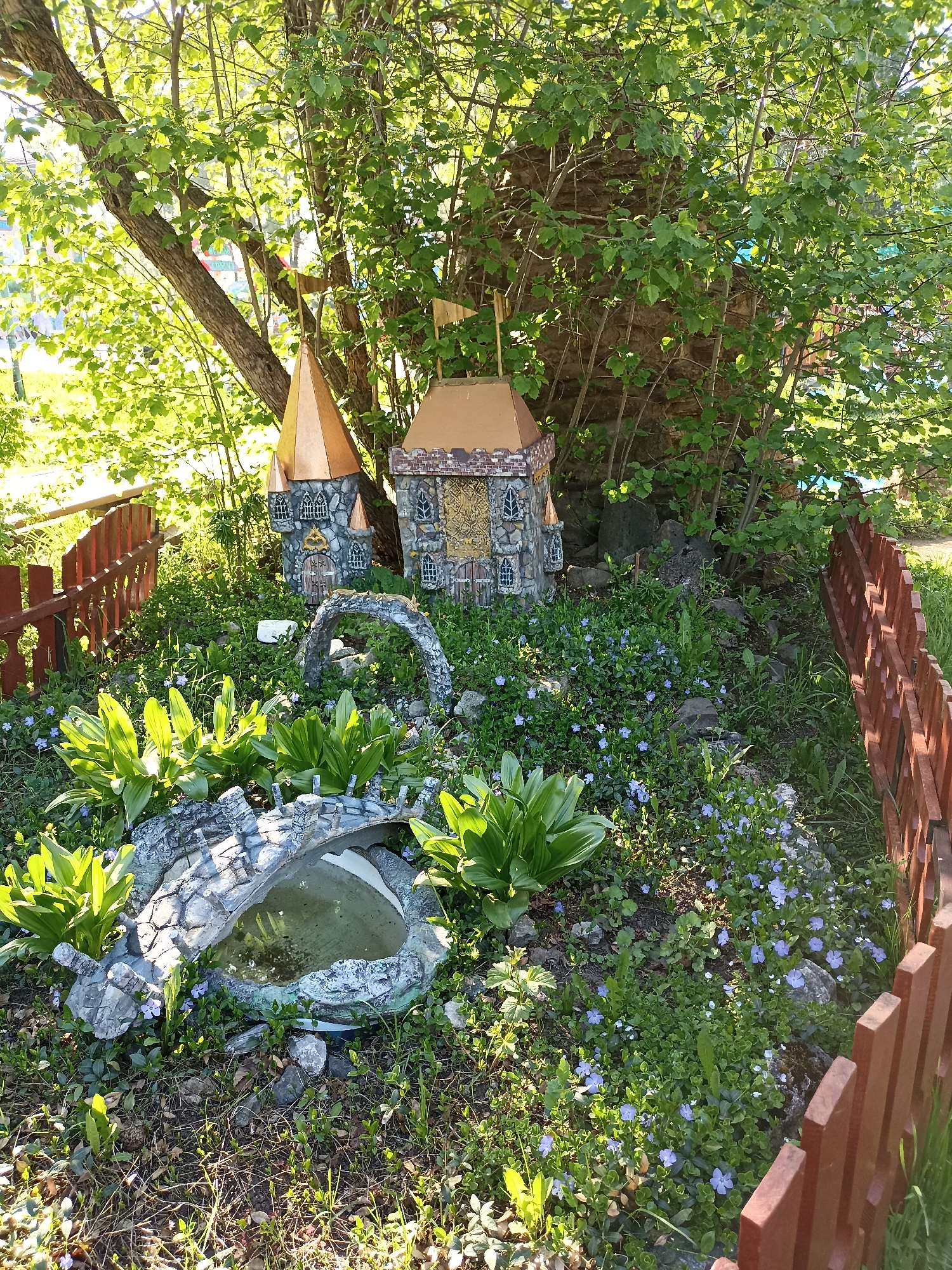 A garden scene in an undisclosed location features two miniature decorative castles situated amongst lush greenery. A small, faux stone bridge spans a pool of water, surrounded by plants with broad, green leaves and scattered purple flowers. The garden is bordered by a low, wooden picket fence. A tree with dense foliage provides partial shade to the area. The light suggests it is daytime, likely in the late morning or early afternoon. No people are visible in the image.