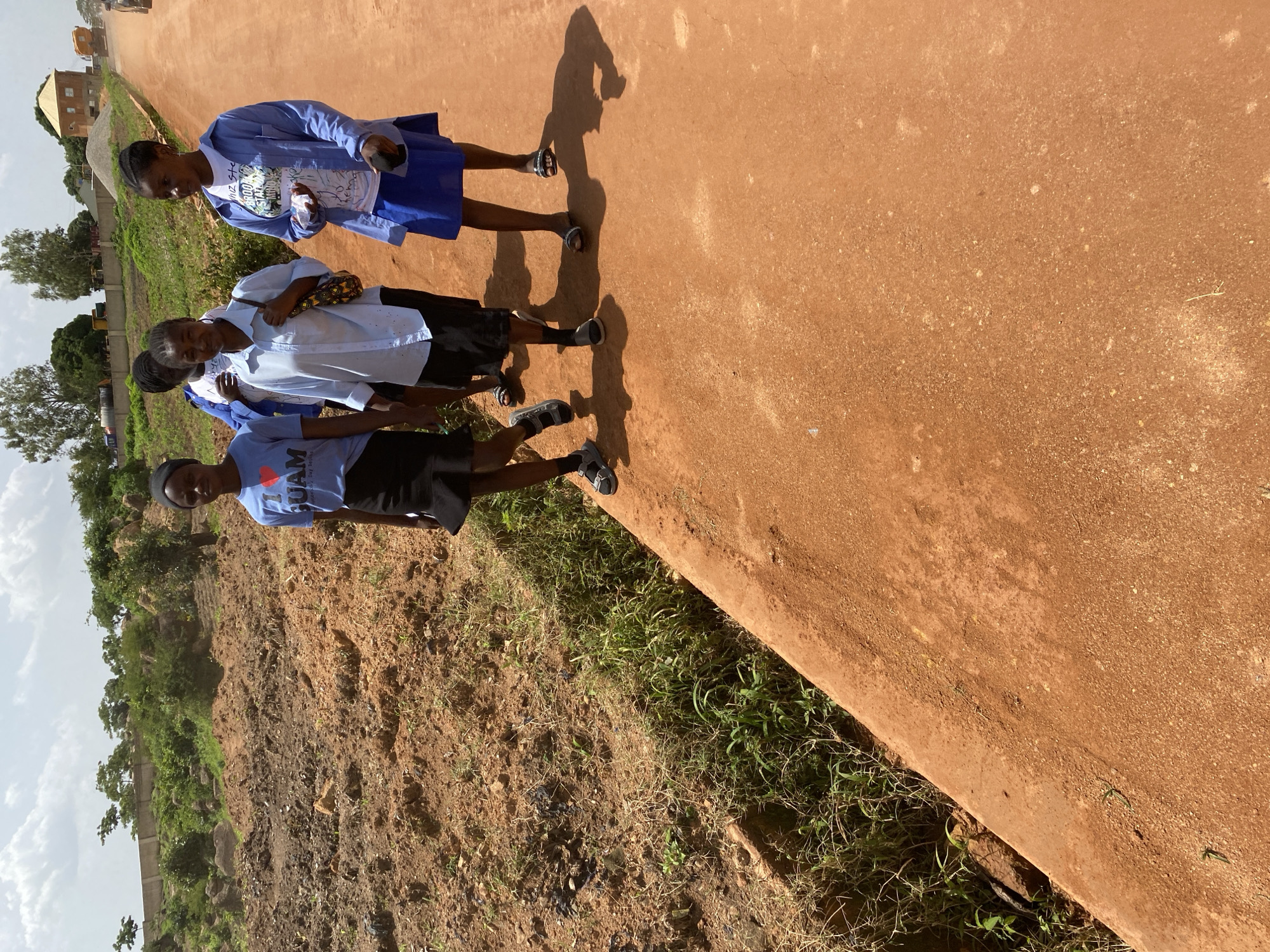 The image shows four young girls walking on a dirt road in Bukuru, Nigeria. The girls are wearing school uniforms, with two of them in blue shirts and skirts, and the other two in light blue shirts and dark skirts. The road is unpaved and appears to be made of red clay. To the left of the road, there is a steep bank with dry grass and some greenery. In the background, there are buildings, trees, and a construction site with a pile of sand and gravel. A truck is visible in the distance, on the road. The sky is cloudy. The girls are walking in the same direction, and their shadows are cast on the road. The overall scene suggests a rural or semi-urban environment in Nigeria.