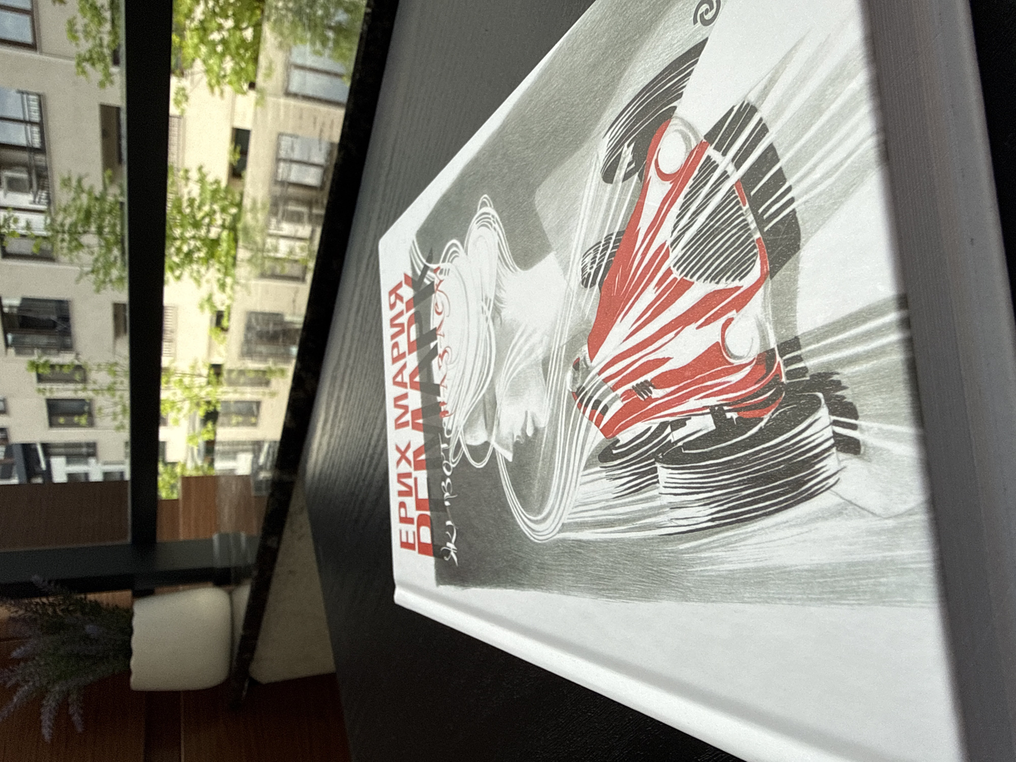 The image depicts a hardcover book resting on a dark surface, likely a table. The book cover features a black and white illustration with red accents, possibly a stylized depiction of a female figure and a race car. Text in red and white is visible along the spine.
Outside a window, a building exterior with several windows is visible, along with green foliage, likely trees. The scene appears to be indoors, near a window with a view of an urban environment. The image was taken in Sofia, Bulgaria.