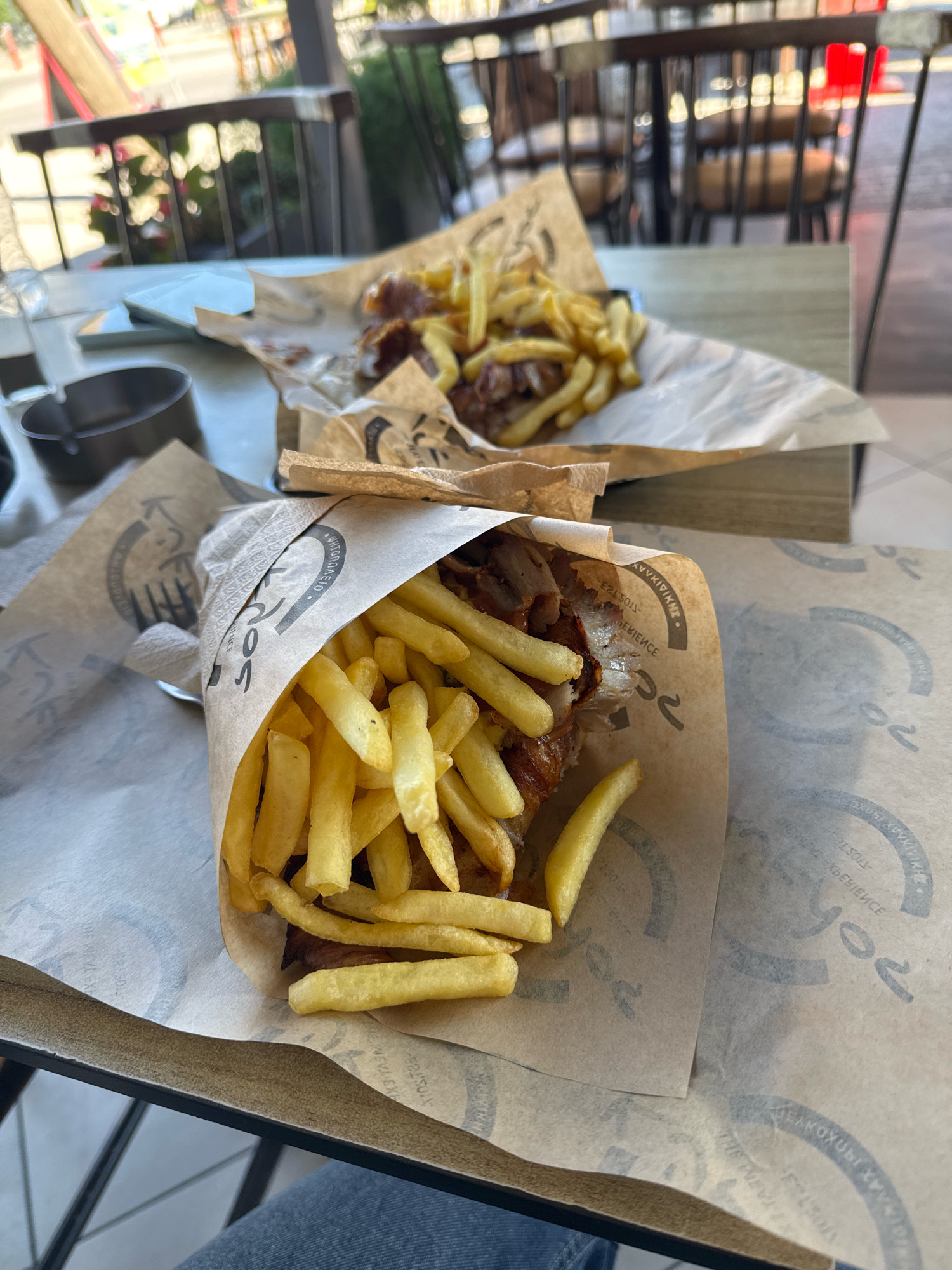 Two servings of gyros and fries are presented on a table at an outdoor restaurant. The gyros are wrapped in paper with Greek lettering and filled with meat and fries. In the background, the restaurant's outdoor seating area is visible with tables and chairs. The scene is lit by natural daylight, suggesting it is daytime. The image does not provide specific information about the city of Pallini Municipal Unit, Greece, beyond the implication that it is an outdoor dining establishment.
