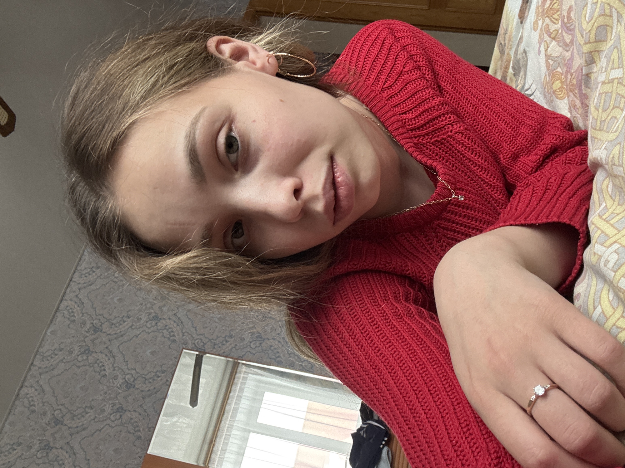 A young woman is laying on her side, presumably in a bedroom or living area. She wears a red, textured sweater, gold hoop earrings, and a delicate gold necklace. A ring adorns her ring finger. A wall with patterned wallpaper is visible in the background, along with a window with blinds. The location may be in Прикамский, Russia, based on the user's request, though nothing in the image directly indicates this.