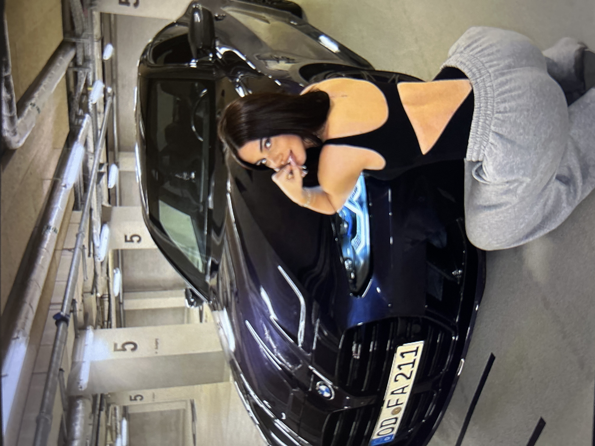 A woman with dark hair is kneeling in front of a dark blue BMW in a parking garage. She is wearing a black bodysuit with a cutout back and gray sweatpants. The car has a German license plate that reads "OD FA 211". The parking garage has concrete pillars with the number "5" stenciled on them and industrial lighting fixtures on the ceiling. The image appears to be taken in a parking garage located in the city of Burgas, Bulgaria.