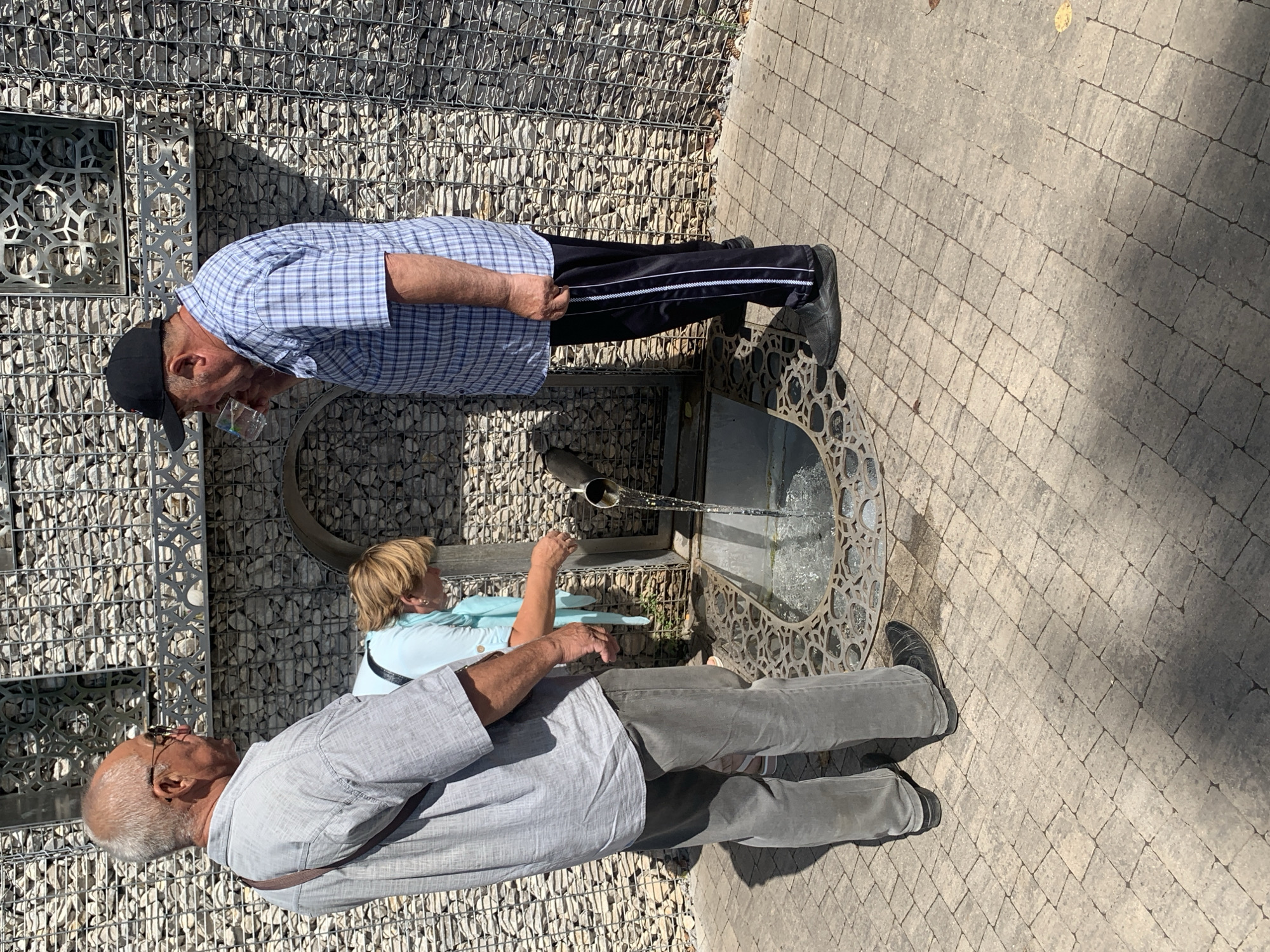 In the city of Arbuzov-Baran, Russia, three individuals are gathered near a public water fountain. The fountain is integrated into a wall constructed of grey stones, with decorative metal elements above. A metal grate covers a pool beneath the spout of the fountain, from which water is flowing.
On the left, an older man with short grey hair and glasses stands facing away from the viewer, wearing a light grey short-sleeved collared shirt and grey trousers. A brown strap is slung across his shoulder.
In the center, a woman with short blonde hair is leaning over the fountain, her back mostly to the viewer. She is wearing a light blue top and appears to be interacting with the water flow.
On the right, another older man, wearing a blue and white plaid short-sleeved shirt, dark trousers with white stripes, and a black baseball cap, is drinking from a clear plastic cup. He is facing towards the fountain.
The ground is paved with light-colored rectangular stones. The overall scene suggests a sunny day, with shadows cast by the people and the wall.