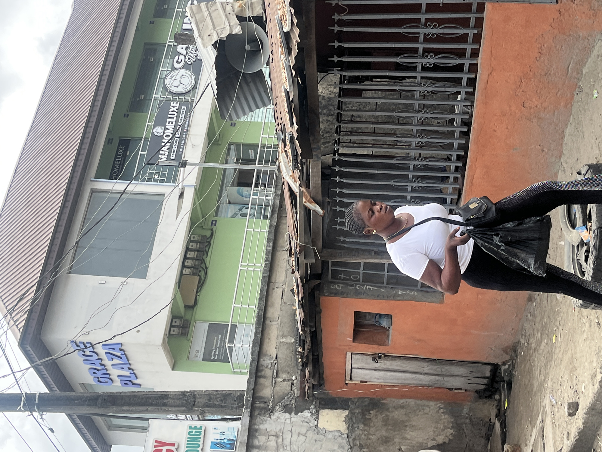 The image shows an outdoor urban scene, likely in Lekki, Nigeria, given the presence of "GRACE PLAZA", "MJANOMELUXE," and the local architectural style. A woman wearing a white t-shirt and black pants walks along a street, carrying a black bag. Buildings are visible in the background, including a multi-story structure with signs for "GRACE PLAZA" and "MJANOMELUXE" and "GA". A megaphone is attached to a corrugated iron structure above the buildings. An adjacent building has an orange exterior wall with a small, weathered door and a barred window above it. Electrical wires crisscross the scene.