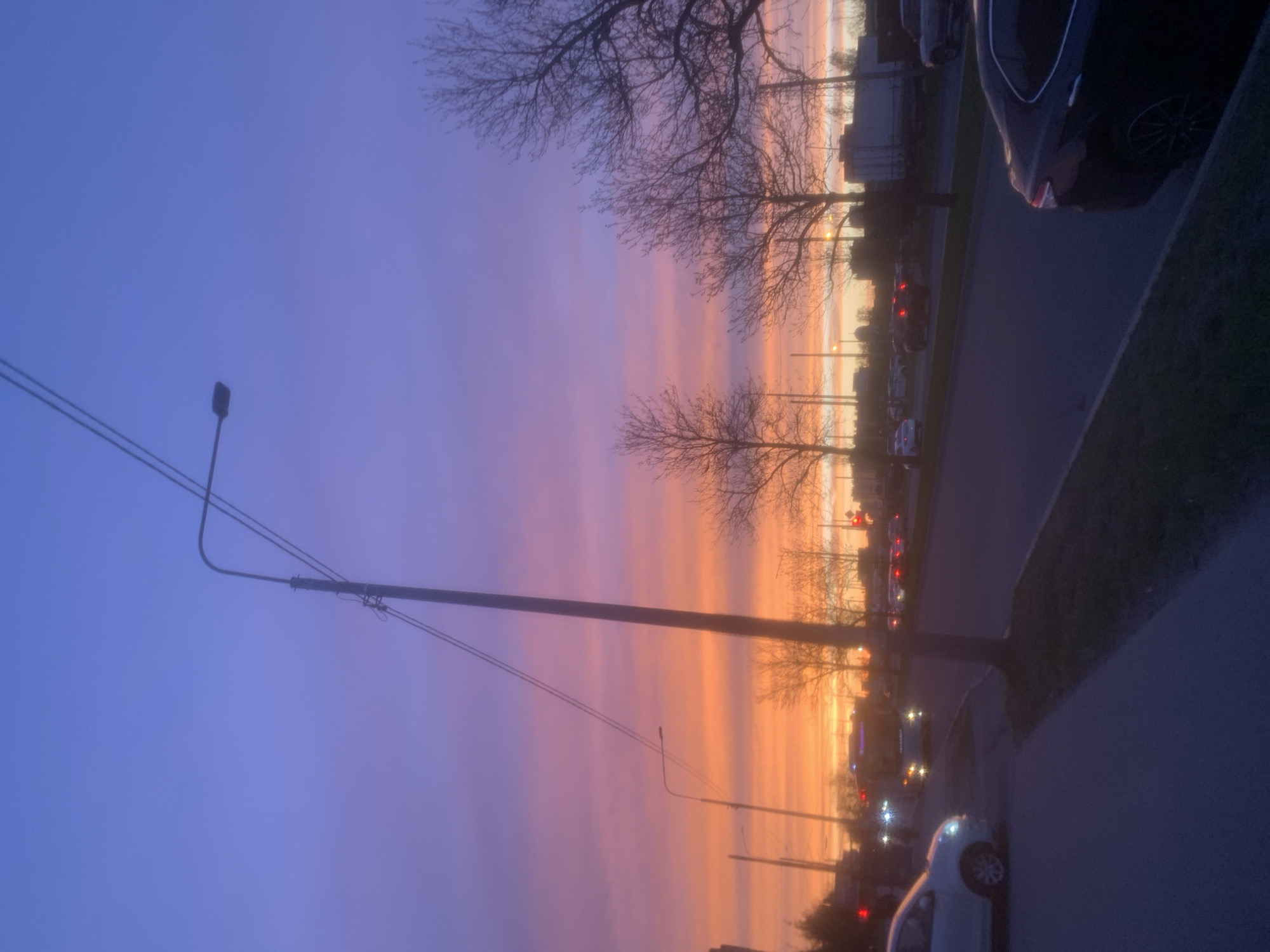 The image captures a street scene at dusk, with a striking sunset dominating the sky. The sky transitions from a deep blue at the top to vibrant hues of orange and pink closer to the horizon. Silhouetted against the sunset are several bare trees and streetlights. A line of parked cars is visible along the side of the road, with one car partially in the foreground on the right side. The scene suggests an urban environment. The location is Unknown, Unknown.
