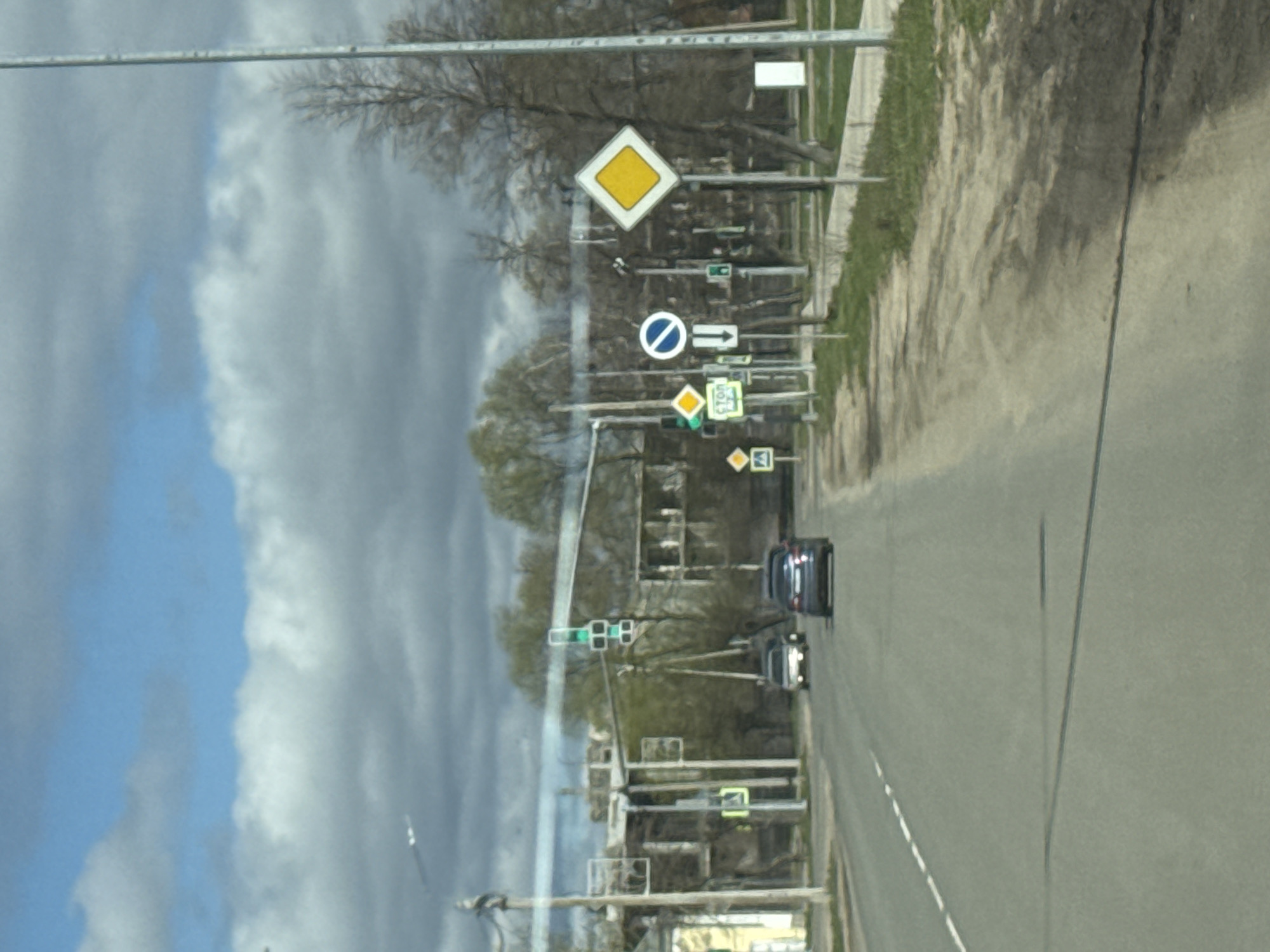 The image shows a street scene in Ново-Никольское, Russia, with a view of a road running alongside a grassy area. Several traffic signs are visible along the roadside, including a yellow diamond-shaped sign indicating a hazard, a blue and white sign indicating "no entry," and other directional and informational signs. Traffic lights are also present. Two cars are seen driving on the road. The sky is visible in the background, showing a mix of blue sky and clouds. The image appears to have been taken from a vehicle.
