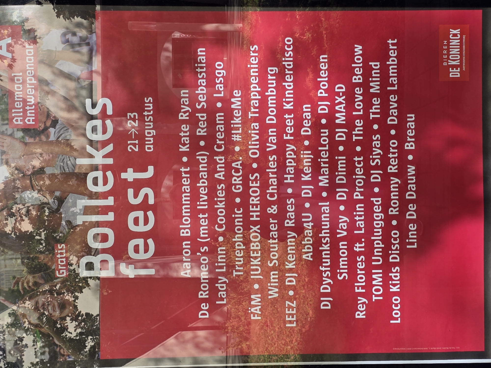 This is a photograph of a red poster advertising an event called "Bollekesfeest" in Antwerp, Belgium. The poster indicates that the event is free and will take place from August 21st to 23rd. A list of performers, including bands and DJs, is featured on the poster. At the bottom right corner, there is a logo for "Bieren De Koninck," an Antwerp-based brewery, with the text "ANTWERPSE STADSBROUWERIJ" below it. The poster is partially obscured by reflections of trees and people, suggesting it is displayed outdoors or behind glass. The background of the poster shows a blurred image of people with their hands raised, common at a festival or concert. The text on the poster is in Dutch.