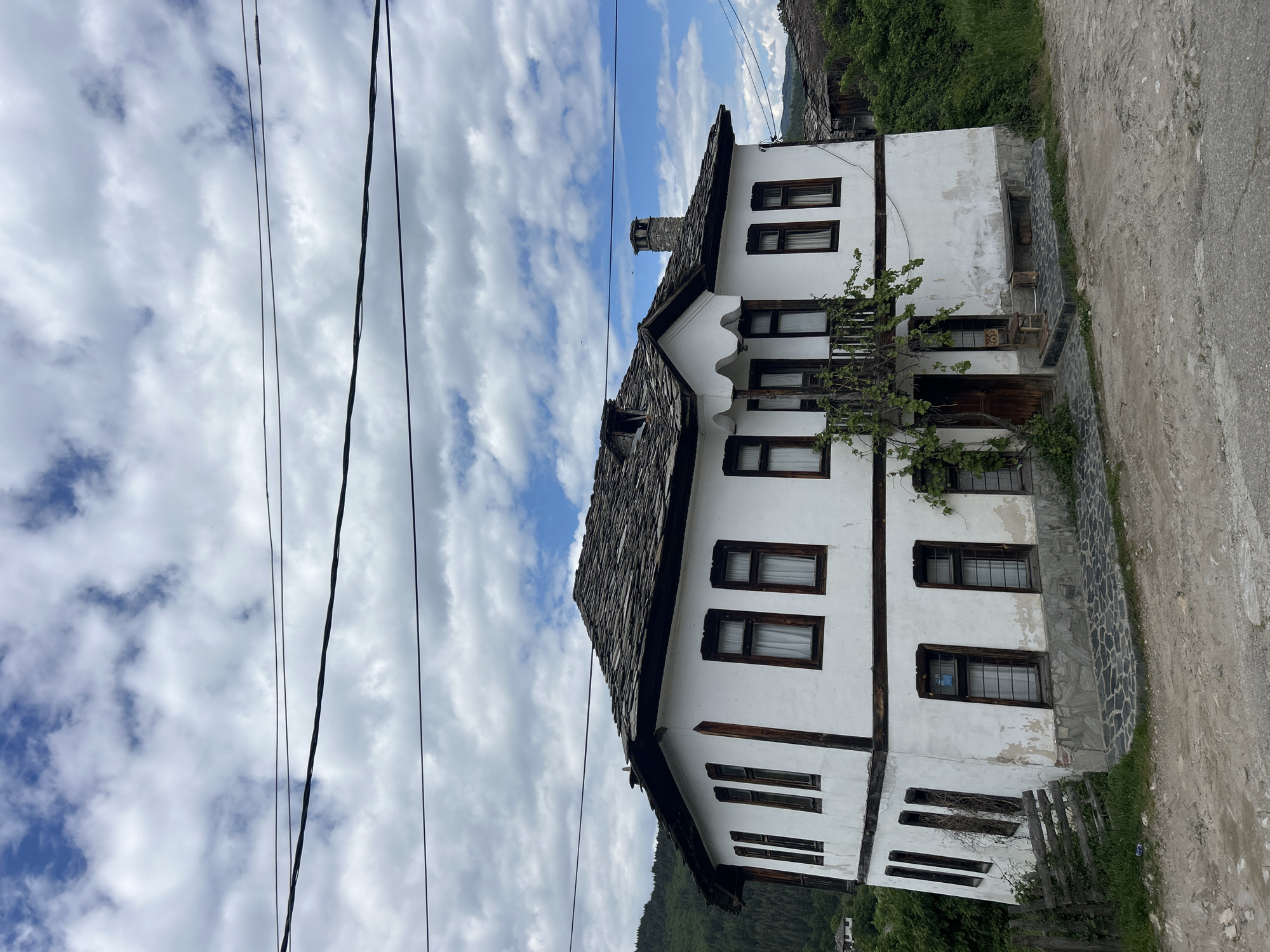 The image depicts a two-story traditional house in the village of Dolen, Bulgaria. The house is white with dark brown wooden window frames and a dark gray stone tiled roof and chimney. A vine is growing near the front entrance on the first floor. Power lines run across the top left of the image, with a sky filled with scattered clouds above. A gravel road runs along the right of the house. A mountain covered with greenery is visible in the background behind the house.