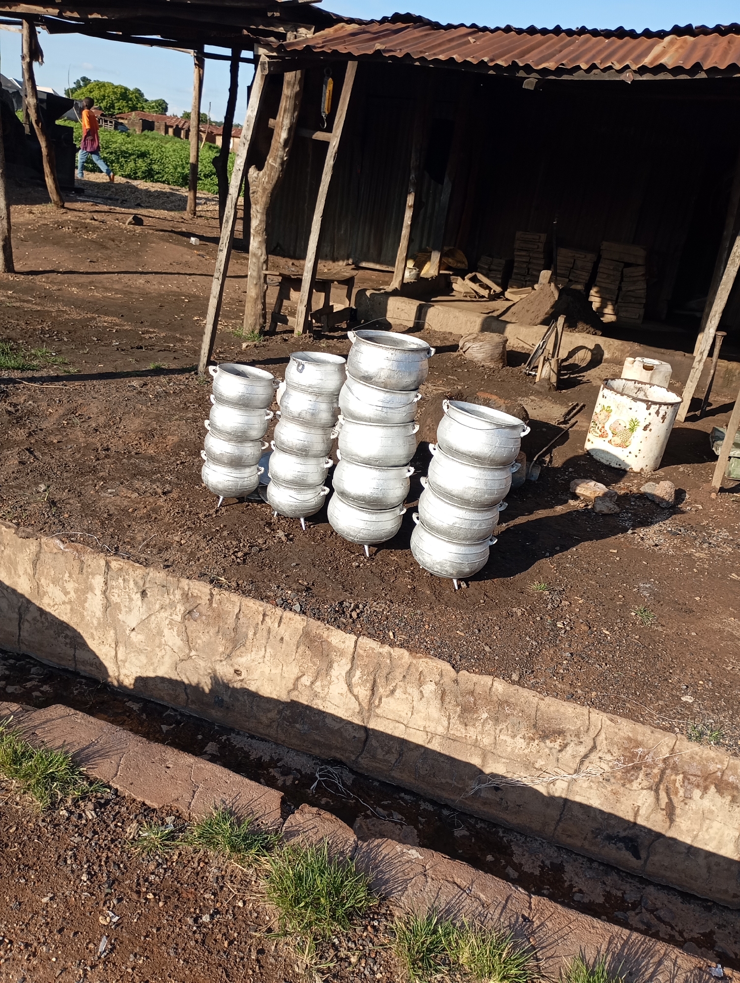 In Shendam, Nigeria, a scene unfolds with a person in an orange shirt and blue pants walking past a rustic outdoor structure. The structure features a corrugated metal roof and wooden supports, suggesting a marketplace or storage area. In the foreground, a row of stacked aluminum pots sits on the dry, earthy ground. To the right, a large, patterned metal container stands, also appearing to be for storage or cooking. The ground is a mix of dirt and gravel, with patches of green grass emerging in places. A concrete structure runs along the bottom of the frame, with a shallow channel of water at its base. The environment appears to be outdoors, with evidence of a sunny day due to the distinct shadows cast by the objects.