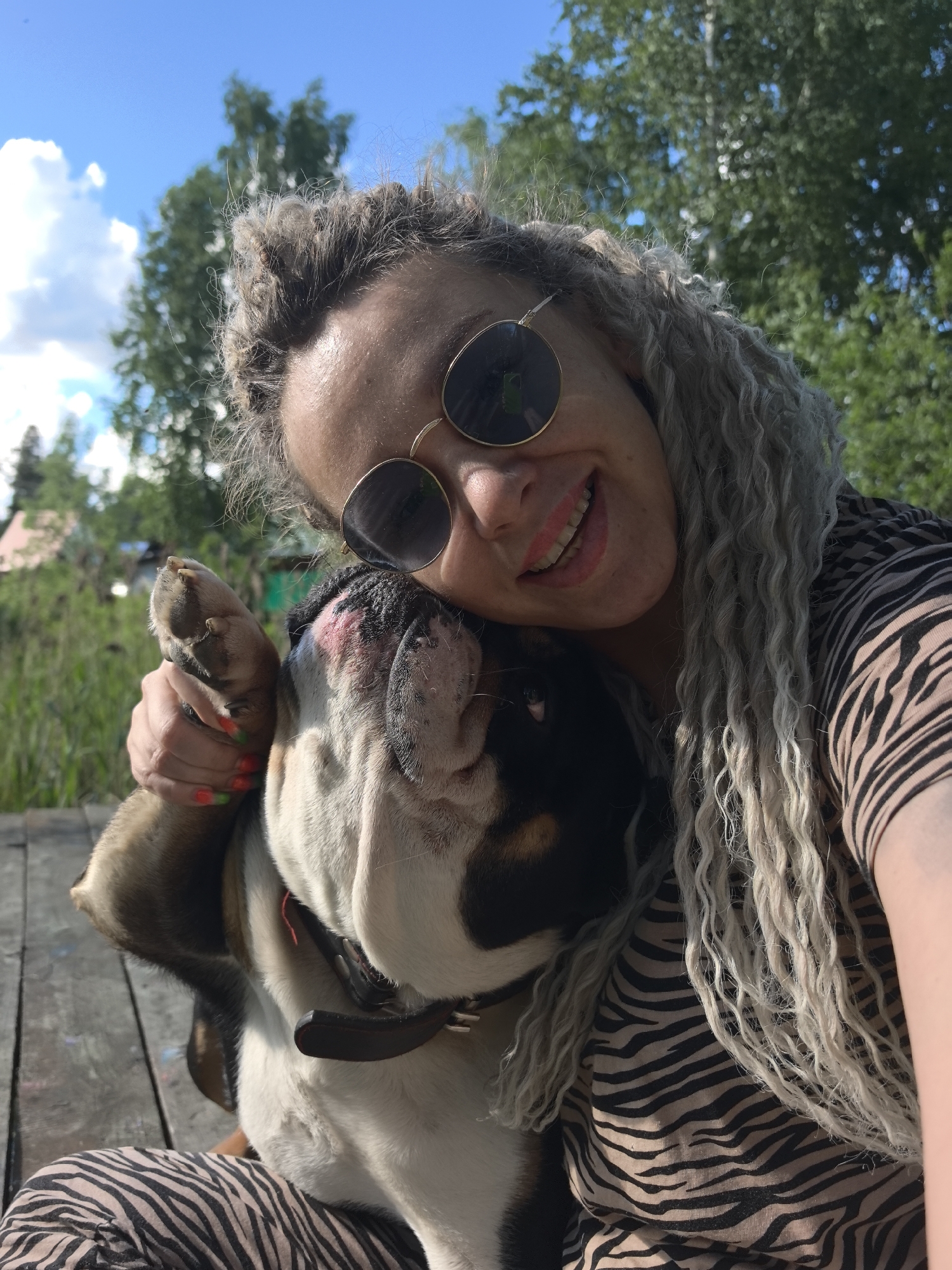 A woman with blonde dreadlocks and round sunglasses is embracing a bulldog. The woman is wearing a zebra-striped shirt and matching pants. She is smiling and has her hand on the dog's paw. The dog has a black and white coat and a brown leather collar. They are sitting on a wooden surface outdoors. The background includes green trees, a blue sky with white clouds, and glimpses of a building. The location is Noviy, Russia.