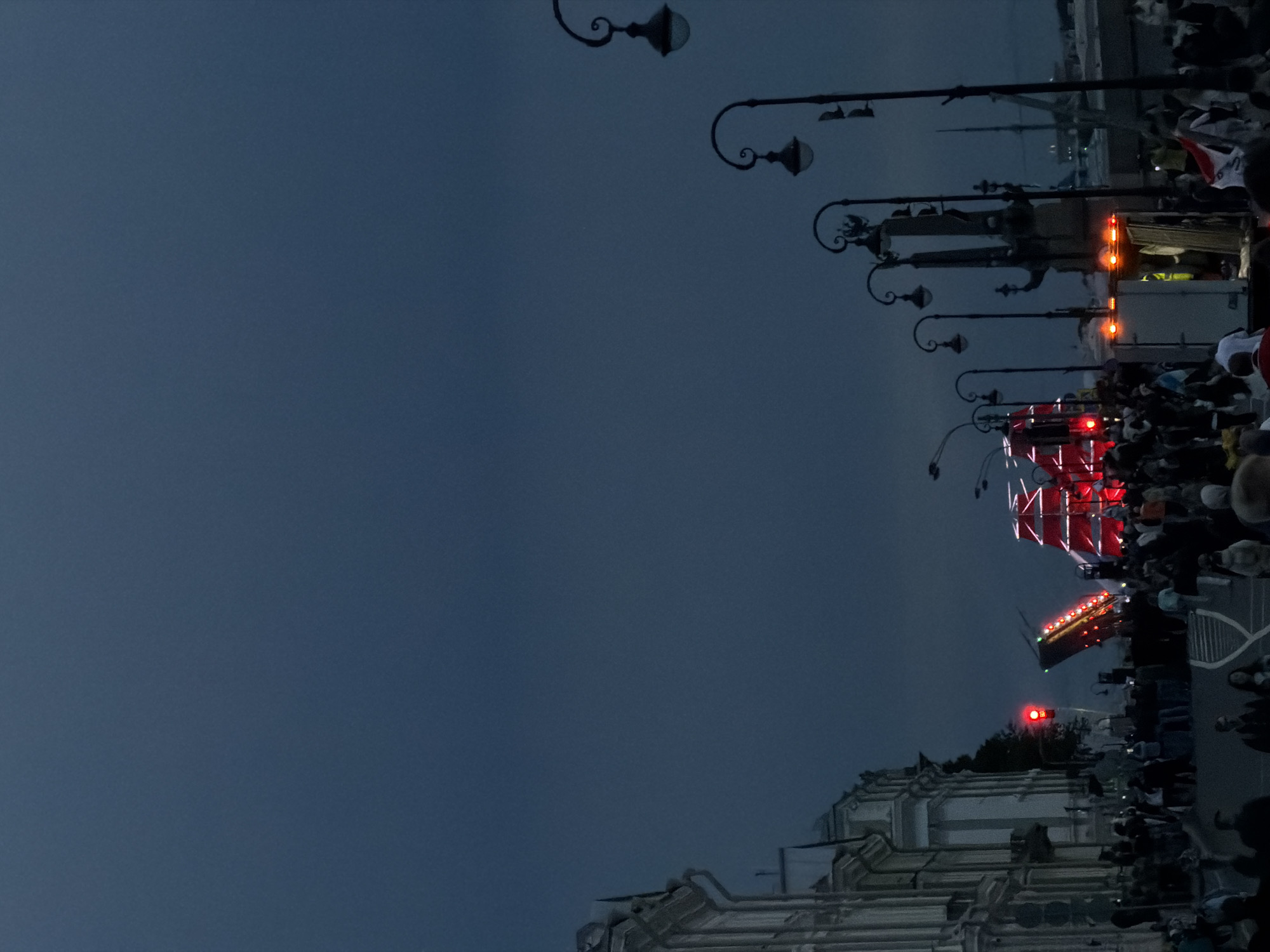 This image captures a nighttime scene in Saint Petersburg, Russia, likely during an outdoor event or procession. The sky is a deep, dusky blue, suggesting either early evening or late night. Ornate street lamps with curved arms and globe-shaped fixtures line the right side of the frame, casting a faint glow.
The main focus of the image is a moving procession or parade. A bright red, illuminated structure, possibly a float or stage, is prominent. It appears to be decorated with numerous red lights and possibly white elements. Behind this, a white truck with flashing orange lights is visible, suggesting it's part of the procession or providing support. A crowd of people is gathered along the sides of the street, watching the event. They are mostly silhouettes against the dim light, but some details of their clothing and presence are discernible.
Further down the procession, another brightly lit structure, also red, is visible, and beyond that, what appears to be a red traffic signal adds another point of light. The architecture in the foreground on the bottom right suggests an urban setting, possibly with grand buildings or facades. The overall atmosphere seems to be one of public gathering and observation, with the lighting and the movement of the procession indicating a lively event. No discernible text is visible in the image.