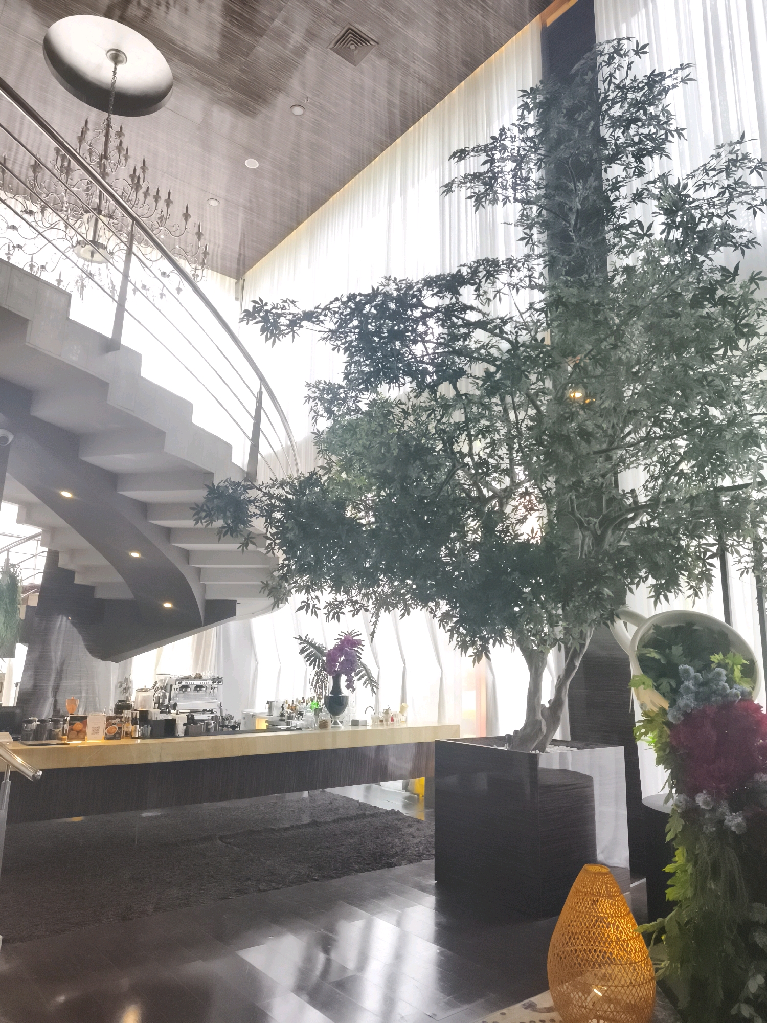This is an interior shot of a modern establishment, likely a cafe or restaurant, located in Palembang, Indonesia. The scene is dominated by a large, artificial tree positioned in the foreground. Behind the tree, a white, sheer curtain forms a backdrop, allowing diffused light to enter the space.
To the left of the tree, a sweeping, curved staircase with a metal railing ascends, leading to an upper level that is not fully visible. The ceiling features a wood-paneled finish, recessed lighting, and a decorative chandelier.
In the mid-ground, a polished wooden counter serves as a bar or serving area, equipped with coffee machines and various bottles. A dark, textured rug lies on the floor in front of the counter.
The overall ambiance is one of contemporary elegance, with a blend of natural and artificial elements. No people are visible in the image, and no specific actions are being depicted. The lighting suggests it is daytime.