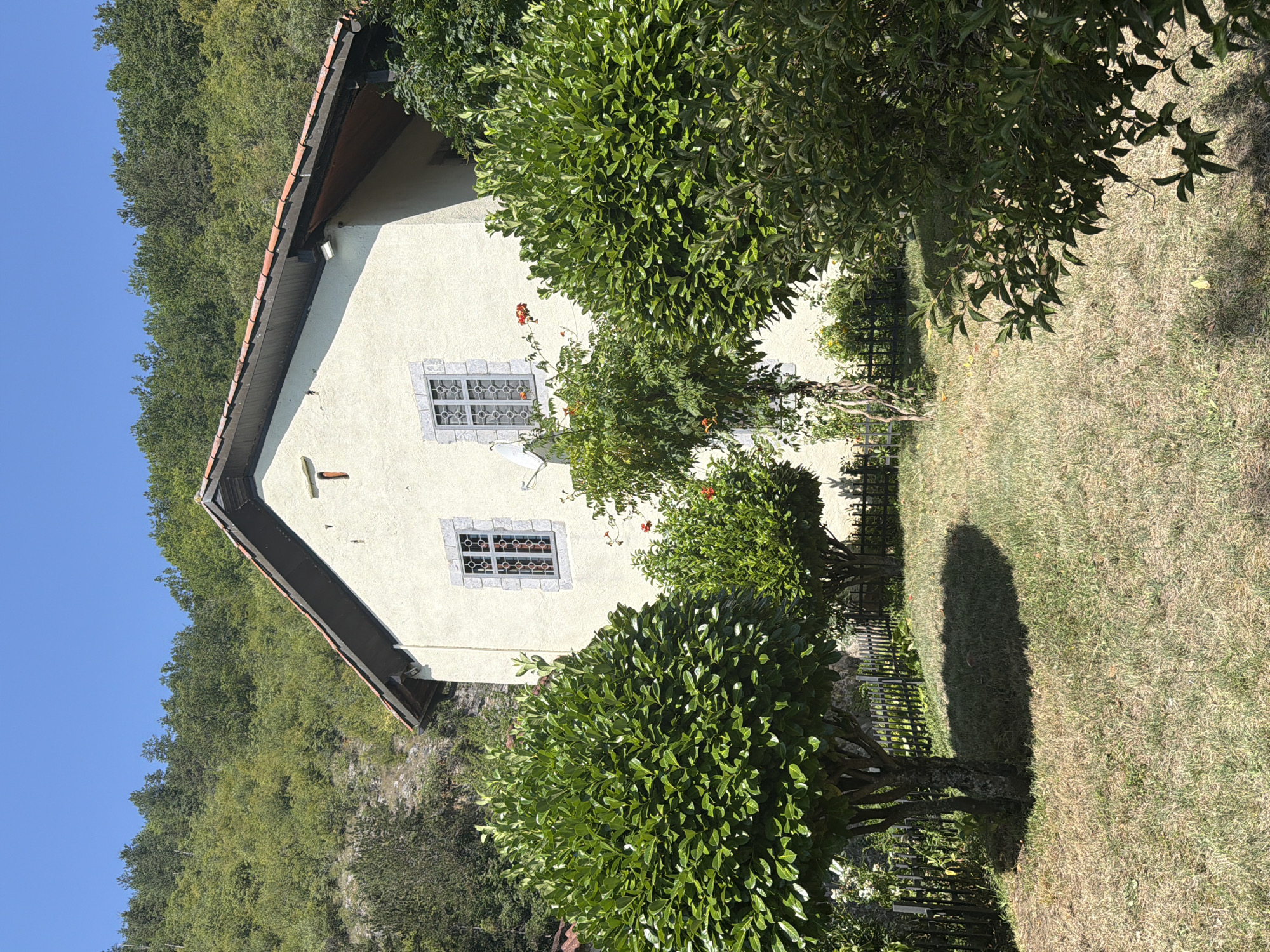 The image shows a building in Manastir Morača, Montenegro, situated on a hillside. The building is light-colored with a red tiled roof and features two windows with decorative grates. In front of the building, there are several trees and bushes, with a lawn in the foreground. The background consists of a densely wooded hill under a clear blue sky. The scene appears to be quiet and serene, with no people or animals visible. The time of day is likely midday, given the bright sunlight casting shadows on the lawn.