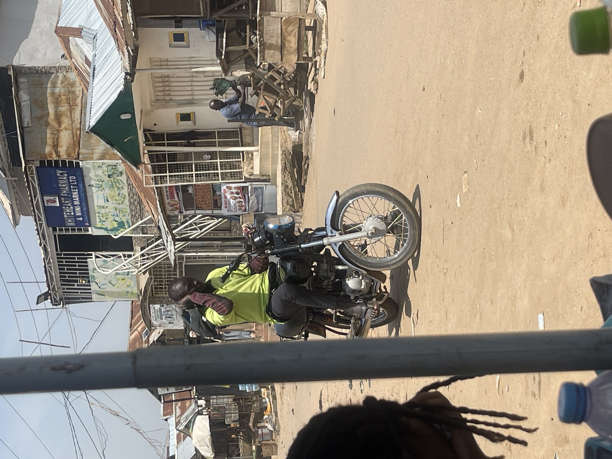 The image depicts a street scene in Abuja, Nigeria, on what appears to be a sunny day. A man is seated on a motorcycle, wearing a bright green shirt and dark pants, with his gloved hands resting on the handlebars. The motorcycle is positioned in the foreground, with its front wheel and handlebars clearly visible.
Behind the motorcycle, a bustling street is visible with several buildings and market stalls. One building has a sign that reads "WHITEHEART PHARMACY & MINI-MARKET LTD". Another man is seen in the background, appearing to be interacting with a pile of wooden materials. The architecture suggests a typical urban environment in Nigeria, with corrugated iron roofs and open-fronted shops.
The overall impression is one of everyday life unfolding in a busy marketplace or commercial area. The sunlight casts shadows, indicating it is likely daytime. The scene is dynamic, with elements of commerce and transportation coexisting.