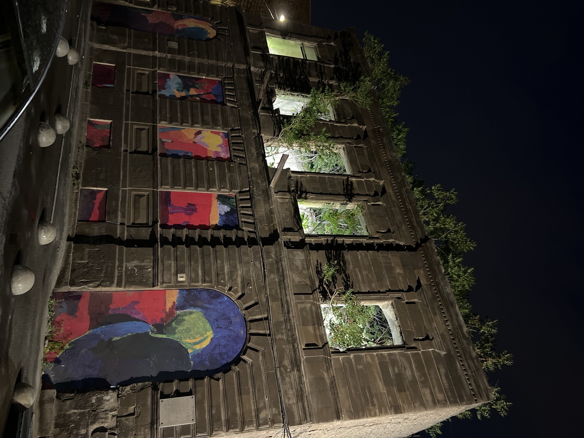 The image depicts a building in Yerevan, Armenia, at night. The building has a distinctive architectural style, with brightly colored abstract paintings decorating its exterior. Vegetation is growing from some of the windows, giving a natural touch to the urban structure. The scene is illuminated by artificial lighting, highlighting the building's facade against the dark night sky. The building appears to be located in a central area of Yerevan. The architectural style is that of the House of Artists after Hakob Kojoyan, known for its facade with mosaic-like paintings.