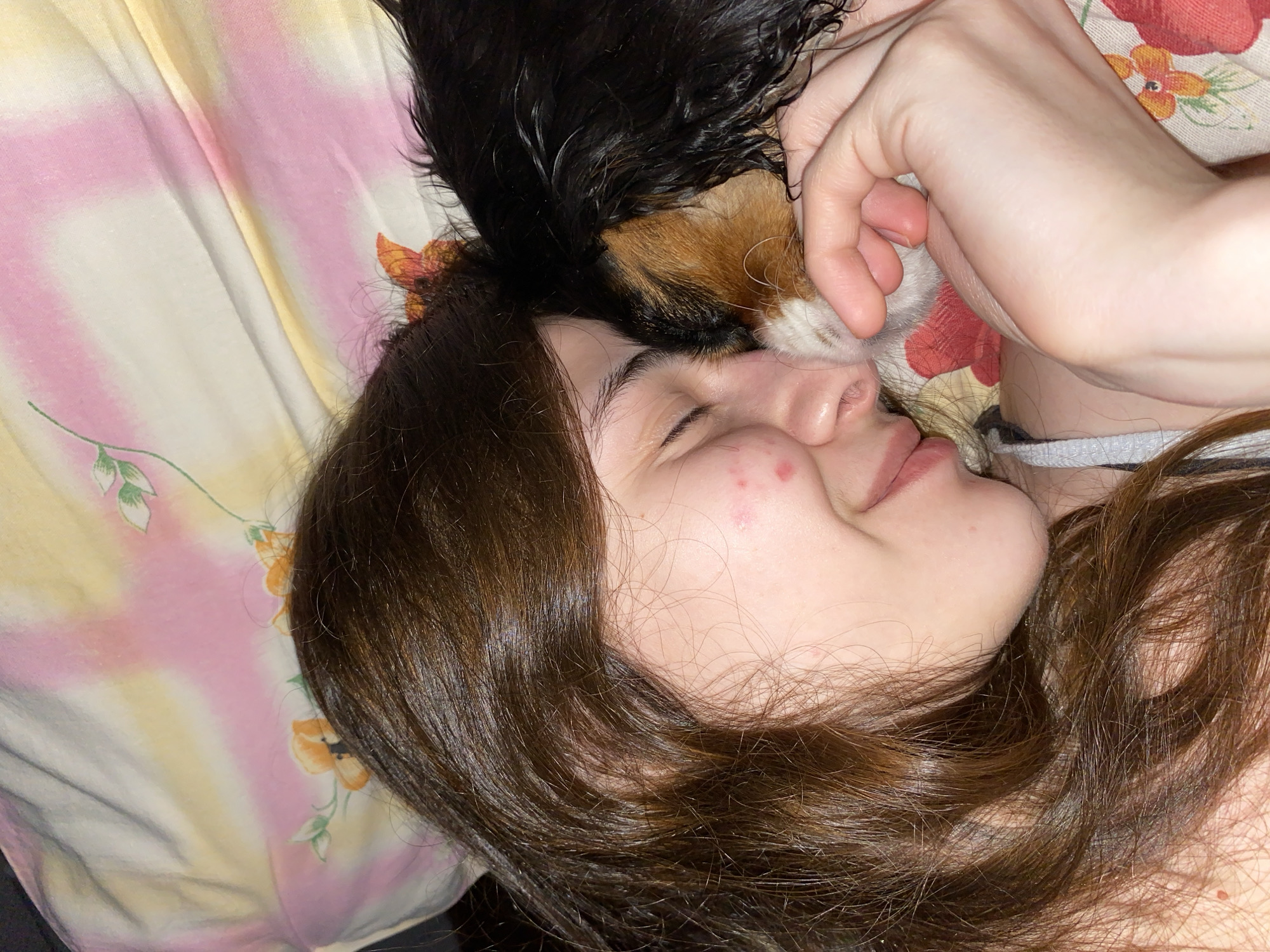 An intimate, close-up shot captures a person and a small dog resting together, likely indoors in Votkinsk, Russia. The person, with long, dark brown hair, is lying on their side with their eyes gently closed or looking downwards, a soft smile gracing their lips, conveying a sense of peace and contentment. A few small red spots are visible on their cheek.
Nestled close to the person's face and head is a small dog, appearing to be a puppy, with black, tan, and white fur. A human hand, likely belonging to the person, is tenderly placed near the dog's head, suggesting a moment of affection and comfort. The pair are resting on bedding or a blanket featuring a vibrant tie-dye pattern of pink, yellow, and white, adorned with floral designs of orange flowers and green leaves. The lighting is soft, contributing to the cozy and relaxed atmosphere of the scene. No visible text is present in the image.