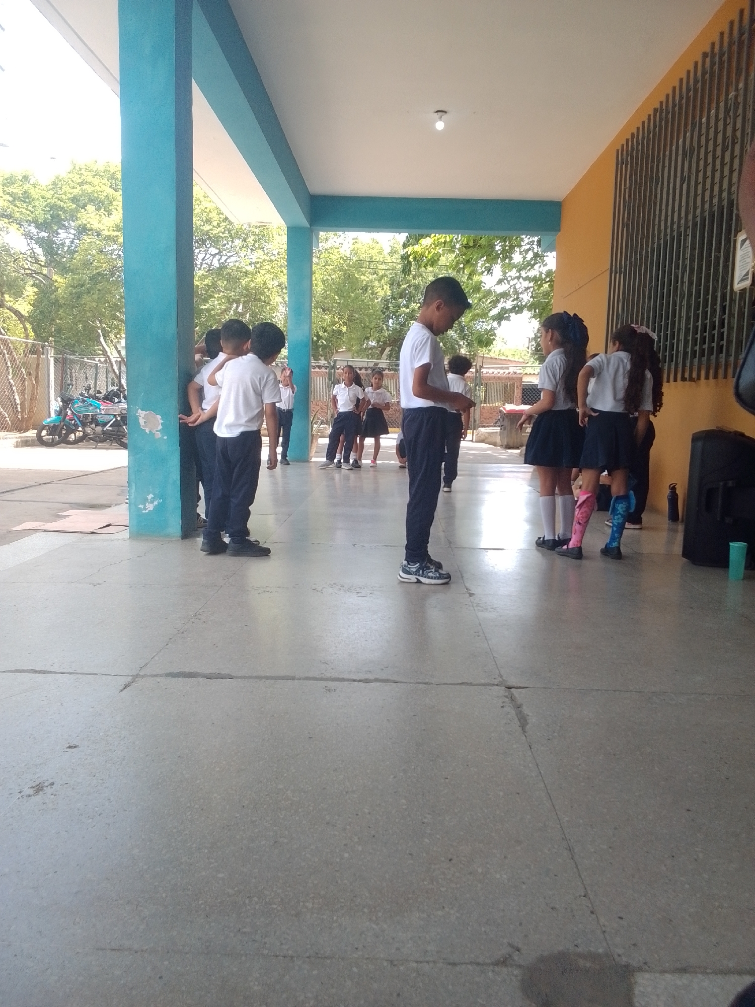 The image shows a group of children, likely students, gathered outside a building. They are dressed in school uniforms, consisting of white shirts and dark pants or skirts. Some of the children are facing away from the camera, while others are looking towards each other or down. The setting appears to be a school or educational facility, with a covered walkway, turquoise pillars, and a yellow building with barred windows. Trees and fencing are visible in the background, suggesting an outdoor environment. The ground is paved with large concrete slabs, and some shadows indicate it is daytime. There are a few objects visible, including a motorcycle parked in the background, a speaker or sound system on the right side of the frame, and a water bottle. The overall mood seems neutral, with no strong emotions displayed by the children. The location context indicates this scene is in La Tagua, Venezuela.