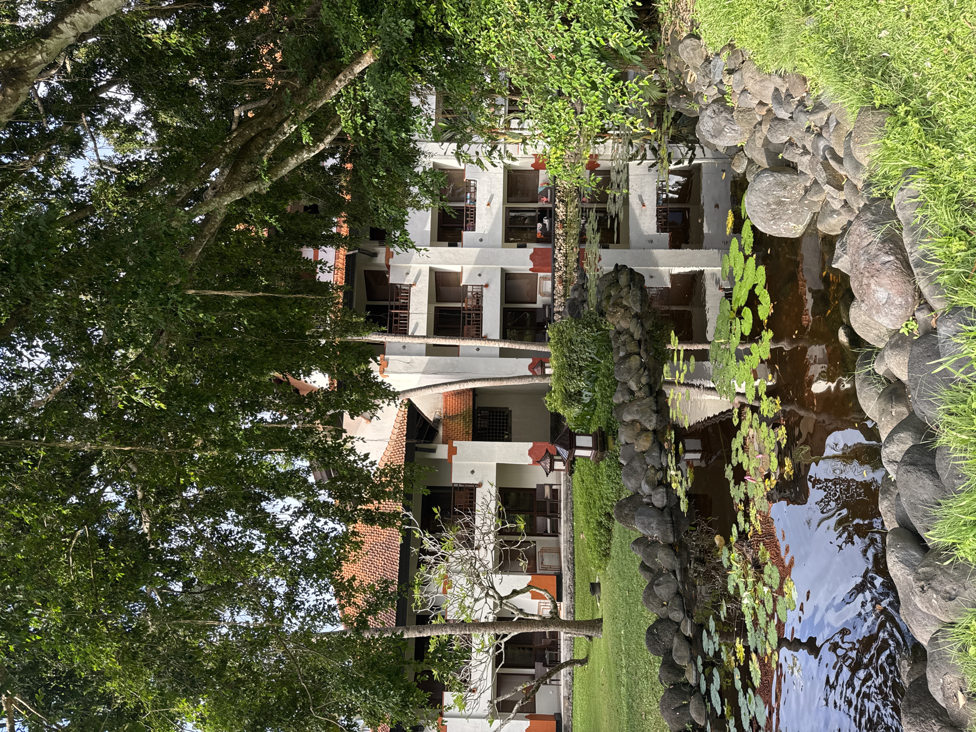 The image shows a building in Kuta Selatan, Indonesia, reflected in a pond. The building is multi-story with a white facade and a red tile roof. Balconies and windows are visible. The pond is bordered by rocks and vegetation, including lily pads. Trees and greenery surround the building, partially obscuring it. The scene appears to be a tranquil garden setting.