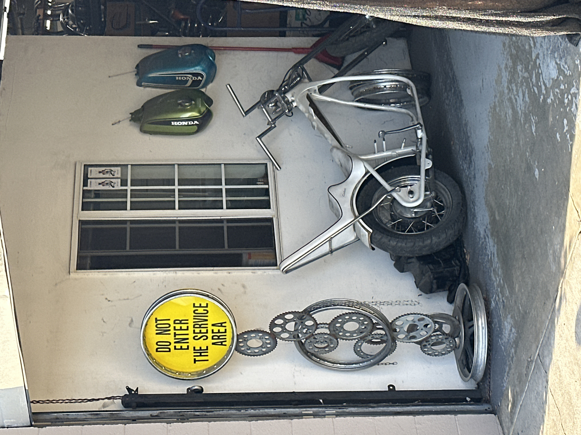 The image shows the exterior wall of a business in Glendale, United States of America. The wall is painted white and features a window. Above the window are two Honda motorcycle gas tanks, one blue and one green. To the right of the window is a disassembled motorcycle frame, and beneath the window, there is a yellow, round sign with black text that reads "DO NOT ENTER THE SERVICE AREA." Adjacent to the sign is a decorative sculpture made from motorcycle gears and a tire rim. Above the wall is a dark area with some objects. The scene appears to be an automotive shop or motorcycle repair shop.