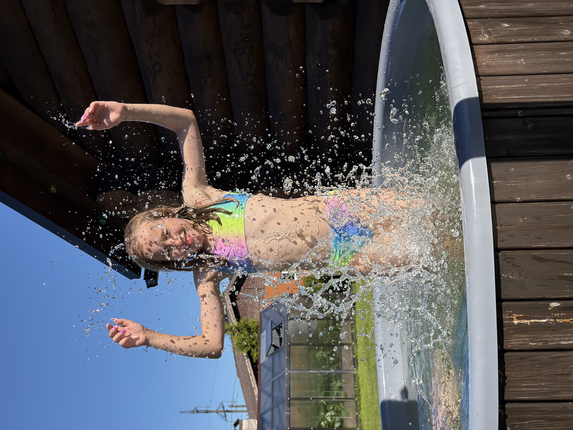 This is a vertically oriented, eye-level, full shot image taken outdoors on a sunny day. The primary subject is a young girl wearing a colorful, tie-dye bikini. She is mid-air, suspended above an inflatable pool, with her arms outstretched and water splashing around her. Her face is tilted upwards, eyes closed, and she appears to be enjoying the moment, a slight smile on her lips. Water droplets are frozen in the air around her, creating a dynamic effect.
The setting appears to be a backyard or a recreational area. Behind the girl, a wooden structure, possibly part of a cabin or a deck, is visible. To the left of the frame, a clear blue sky dominates, with the sun casting shadows and highlighting the water splashes. In the background, a greenhouse and some trees can be seen, suggesting a suburban or rural location. The inflatable pool has a light gray rim and is filled with clear water. The ground around the pool is made of dark wooden planks.
The activity depicted is a playful moment of a child jumping or falling into a pool, creating a dramatic splash. The bright sunlight and clear sky suggest it is daytime, likely mid-afternoon. The overall mood is one of fun and exhilaration. There is no visible text in the image. The location context of Dreiliņi, Latvia, helps to place the scene geographically.