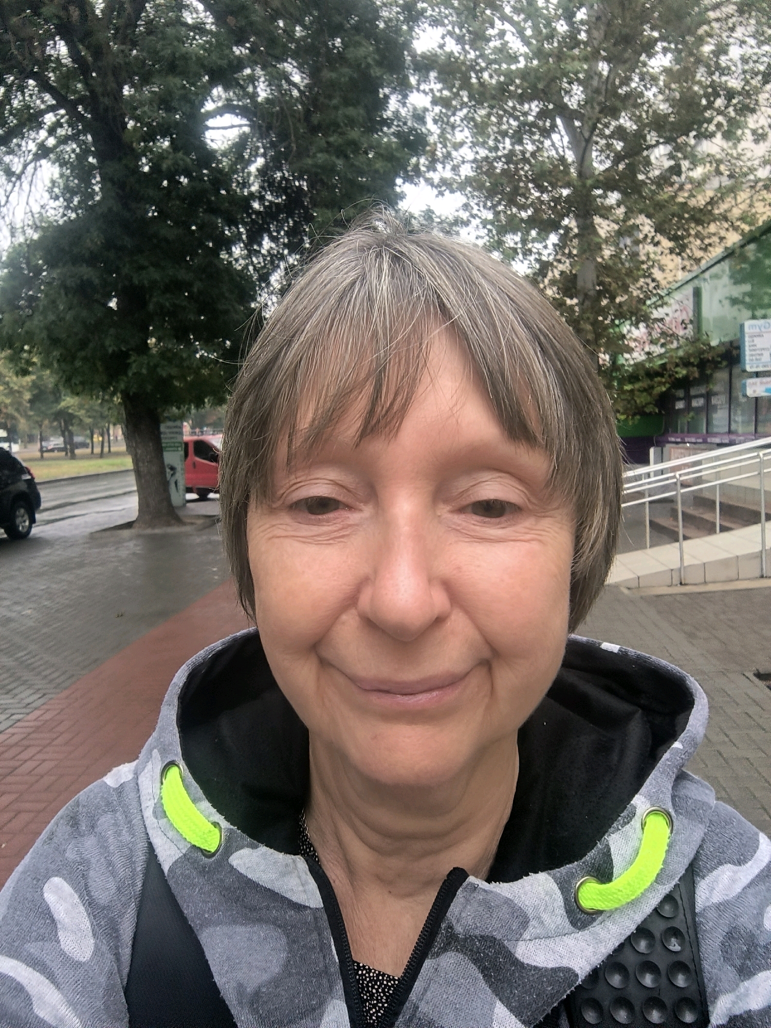 A medium shot selfie captures a woman in Mykolaiv, Ukraine, with her face filling the foreground. She has short, graying hair with bangs and a slight smile. She is wearing a gray camouflage hoodie with bright neon green drawstrings. In the background, a paved sidewalk is visible, appearing wet, suggesting recent rain. To the left, a dark-colored car and a tree are partially visible. Further in the background, more trees, a red van, and buildings line the street, indicating an urban setting. A ramp with railings is visible on the right side of the frame, leading up to a building. The sky is overcast.