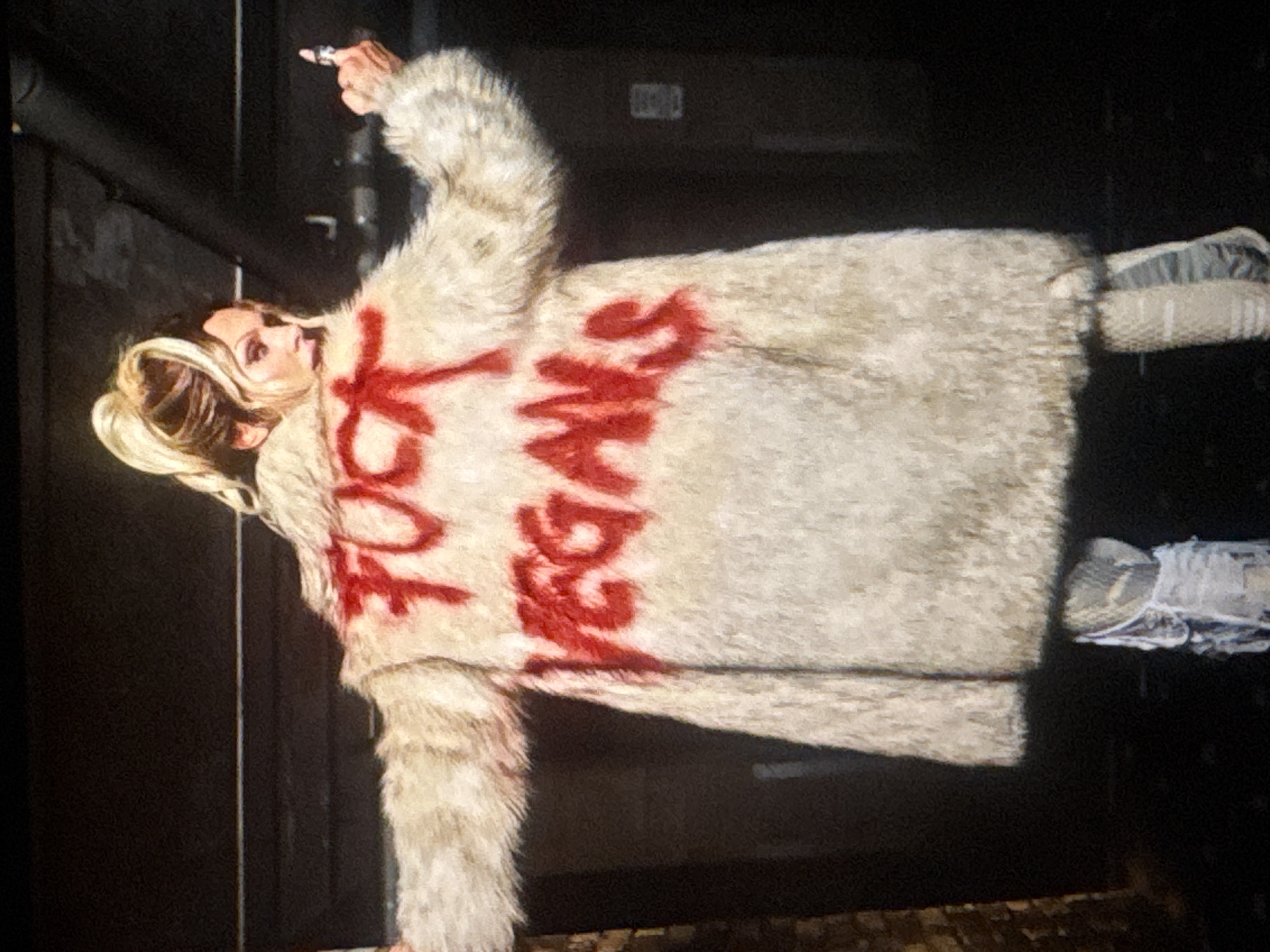The image features a person wearing a fluffy, light-colored coat that appears to be made of faux fur. Red spray paint is used to write text on the front of the coat. The text is partially obscured and difficult to read fully, but some letters are visible. The person is holding what looks like a marker or a pen in their right hand, and they appear to be in the process of writing or adding to the markings on the coat. The person has blonde hair styled up. They are also wearing white sneakers. The background is dark and appears to be an indoor setting with some metallic pipes visible. There is a shadow cast on the right side of the image. The lighting suggests it is not daytime.
