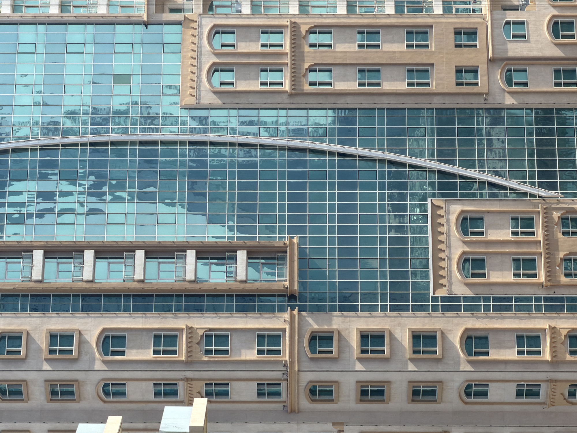 The image captures a close-up, exterior, eye-level view of a modern skyscraper in an urban environment. The building features a striking contrast between its beige, textured facade with decorative architectural elements and its expansive, reflective teal glass windows. The facade has a series of regularly spaced rectangular and arched windows, some with ornate framing. The glass portion of the building is a grid of square panes, with subtle reflections of the sky and possibly other buildings. A prominent vertical silver element curves down the glass facade, adding a dynamic visual line. The composition focuses on the architectural details and textures of the skyscraper, with no people or discernible actions visible. The lighting suggests it is daytime, with sunlight casting shadows on the textured facade.