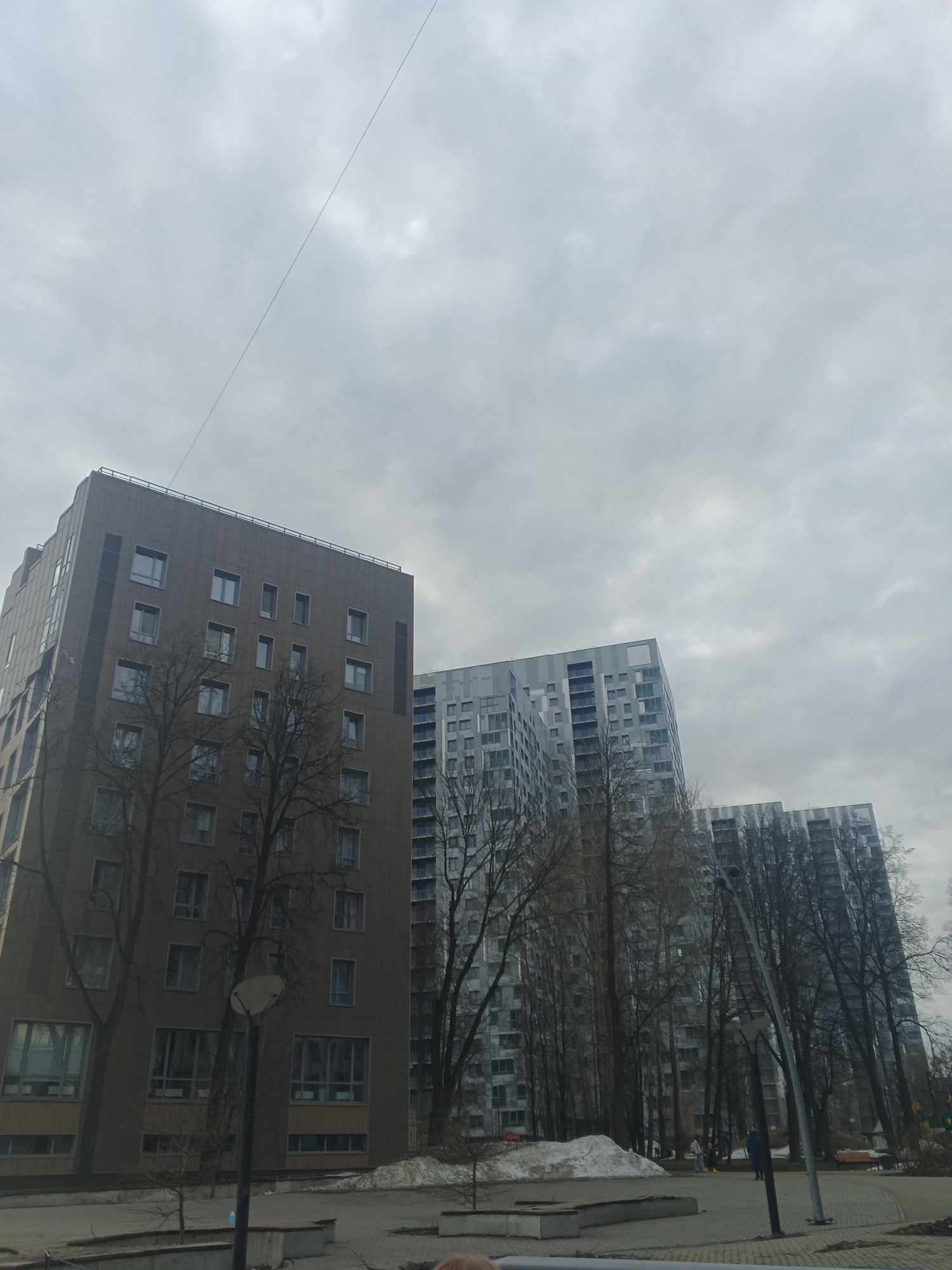 The image shows a cityscape with three high-rise apartment buildings under a cloudy sky. The building on the left is brownish, while the other two are light grey. Bare trees stand in front of the buildings. In the foreground, there is a paved area with benches and patches of snow. A lamppost is also visible. The location is in the city of Perm, Russia.