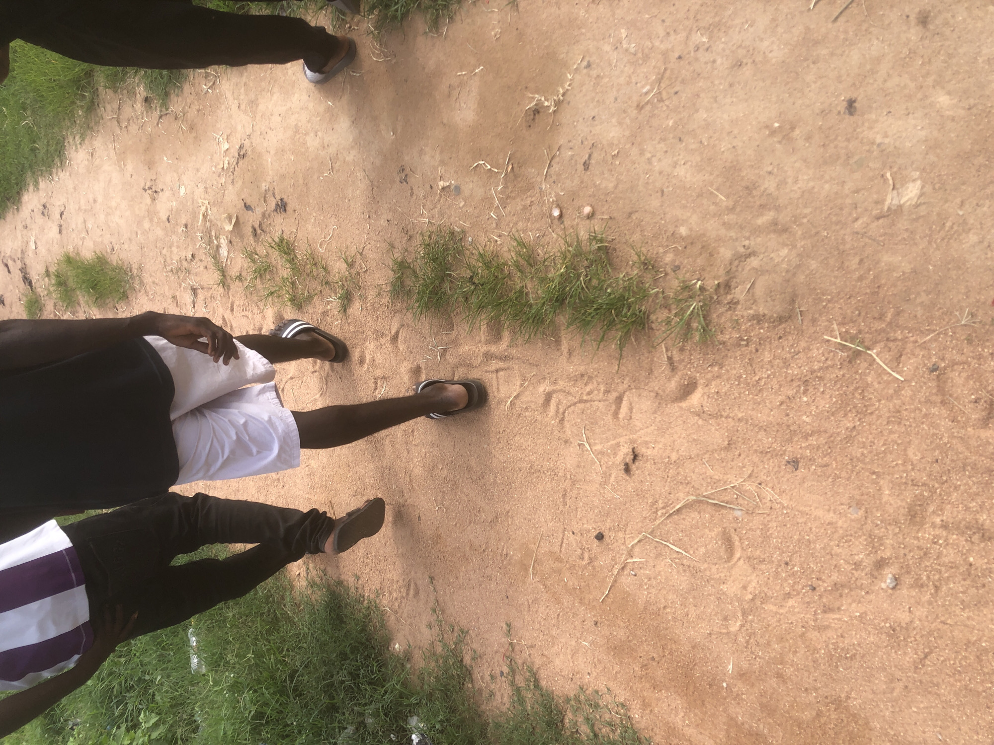 The media file depicts a ground-level view of people's legs and feet on a dirt path in Jalingo, Nigeria. Three individuals are partially visible, their legs clad in casual attire: one in dark pants and flip-flops, another in white shorts and flip-flops, and a third in dark pants and striped shirt with sandals. The path is sandy with patches of dry grass and sparse vegetation. The lighting suggests it is daytime. The scene appears to be outdoors, possibly a rural or unpaved area. The focus is on the lower bodies of the individuals and the texture of the ground, with no discernible text or specific event being captured beyond people walking.