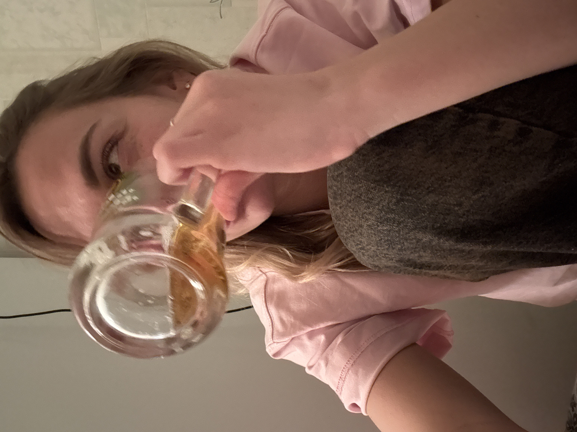 A woman is drinking from a glass mug. She is wearing a pink shirt and dark-colored pants. The background is a plain wall. There is no indication of the location being Ust-Ilimsk, Russia.