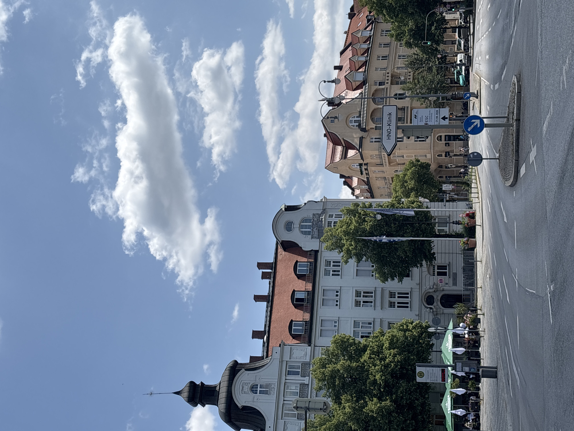 This is a wide-angle, eye-level shot of a street intersection in Munich, Germany, under a partially cloudy blue sky. On the left, a pale gray building with a dark, domed turret topped with a spire dominates the foreground. This building has a mansard roof with red tiles and several dormer windows. To its right, a lighter gray building with a red tiled roof features multiple dormer windows and tall chimneys. Further to the right, a yellow building with a red tiled roof and arched dormer windows is visible. The street in the foreground is paved and marked with white lane lines. A traffic island with a signpost featuring an arrow pointing to "HNO-Klinik" and other information is positioned in the middle of the intersection. Green umbrellas of an outdoor cafe are visible on the left side of the street, with people seated beneath them. Trees with lush green foliage line the street. A traffic light is visible on the right side of the frame, showing a green light. A few vehicles are present on the street, including a turquoise van and several cars. The overall scene depicts a bustling urban environment on a bright, sunny day.