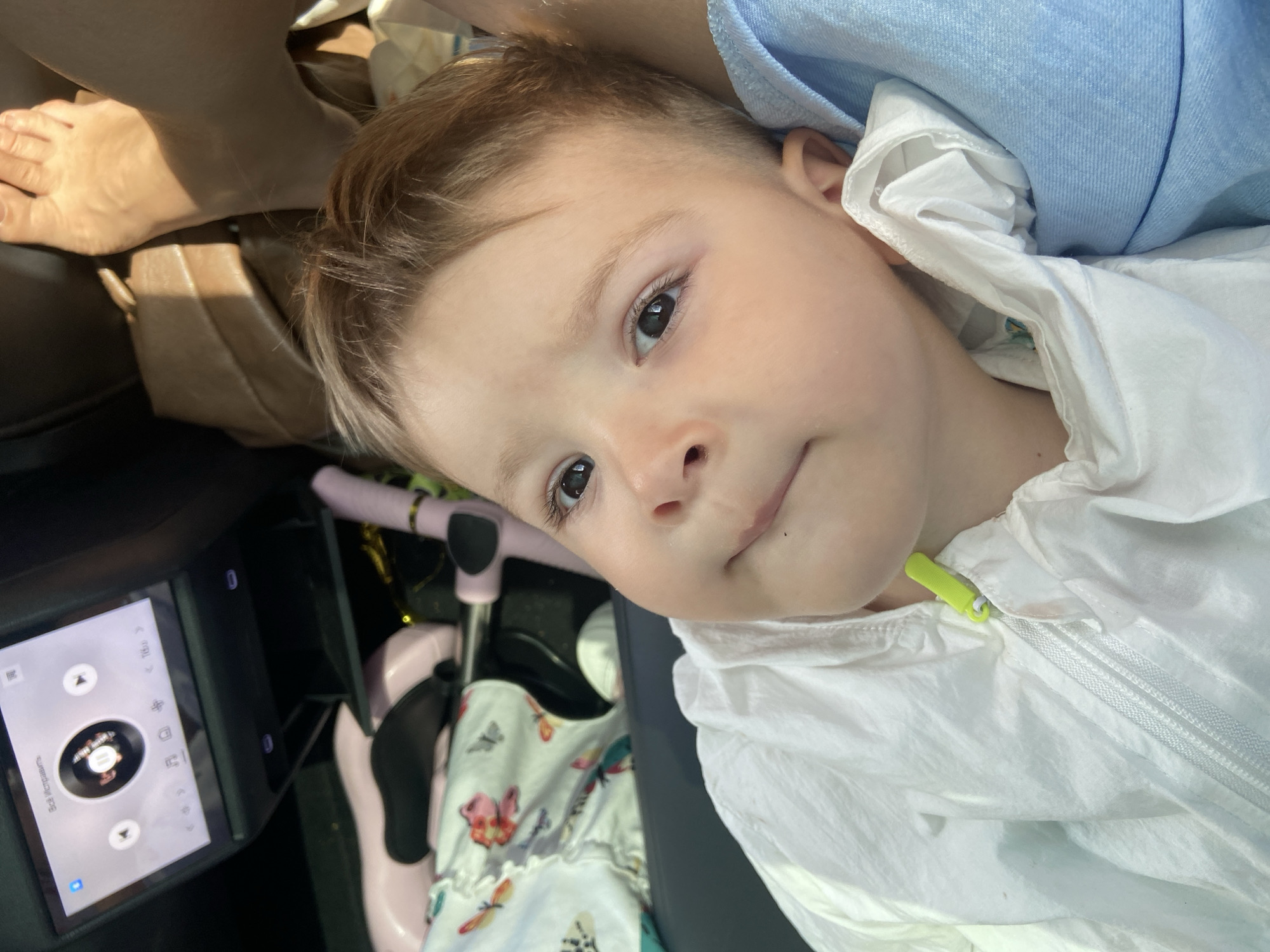 A close-up, high-angle medium shot shows a young child with fair skin, light brown hair, and dark eyes. The child is wearing a white hooded jacket with a neon green zipper pull. The child's head is turned to the left, and they appear to be looking at something off-camera with a slight smirk. The child is nestled against a person wearing a blue garment. In the background, a car's entertainment screen is visible, displaying a music interface with a circular album art. A pink toy, possibly a stroller or similar item, and a white blanket with a floral and butterfly pattern are also present in the background, suggesting a travel or domestic setting within Huangpu District, China. The lighting indicates it is daytime.