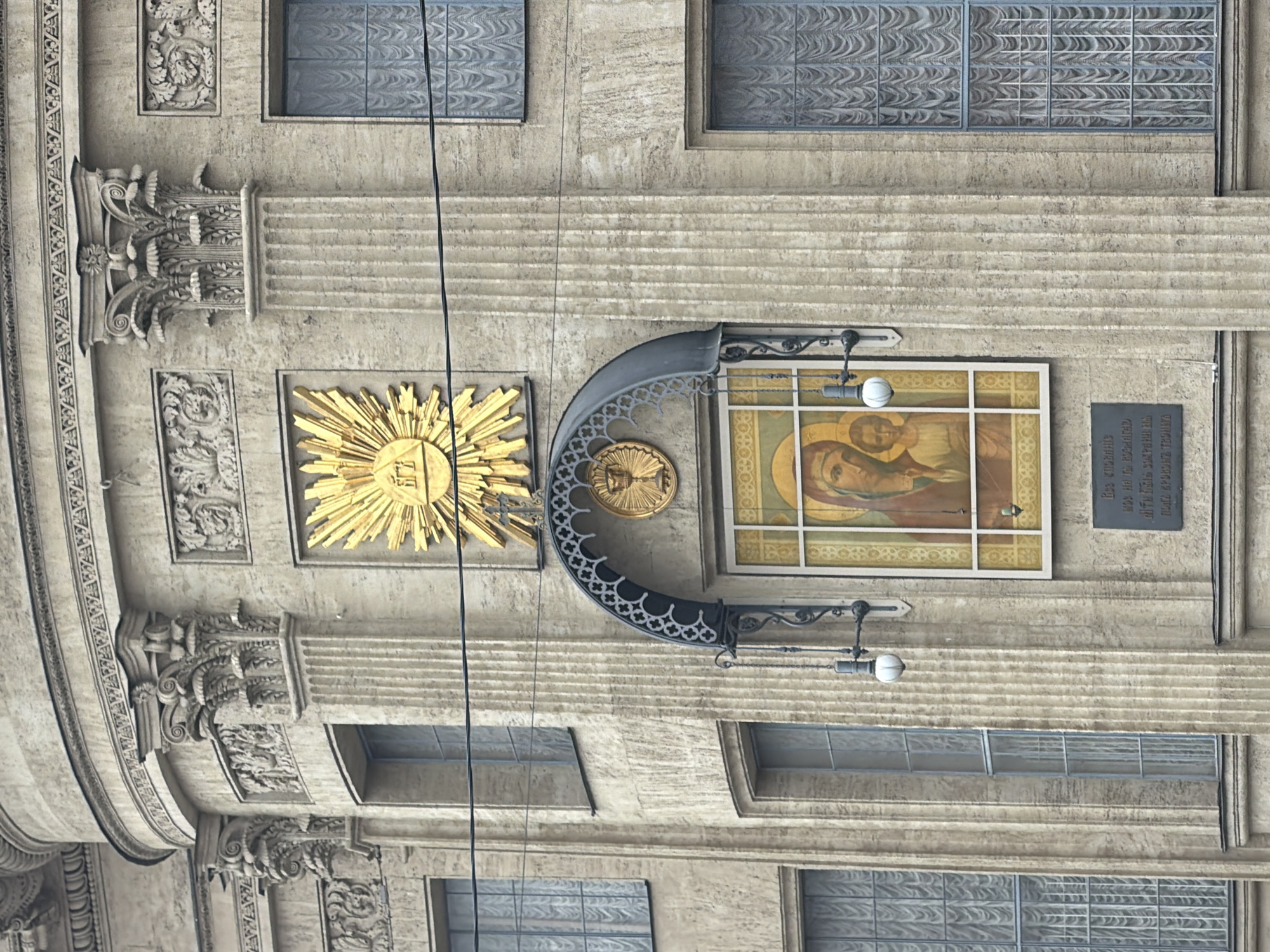 The image displays a detailed section of the upper levels of the Kazan Cathedral facade, located on Nevsky Prospekt in Saint Petersburg, Russia. The building is constructed from a light-colored, textured stone, exhibiting classical architectural elements. Visible features include fluted pilasters or columns topped with ornate Corinthian-style capitals and decorative friezes above them.
Centered on the facade, a large golden emblem shaped like a sunburst contains a triangular motif enclosing a stylized Eye of Providence and what appears to be Cyrillic letters "БГ". Directly beneath this, a prominent rectangular icon depicting the Virgin Mary holding the infant Jesus (Our Lady of Kazan) is recessed into the wall. This icon is framed and situated under a dark, arched metal canopy featuring intricate scrollwork. A small golden circular emblem, depicting a chalice with a cross, is centered within the arch of this canopy. A white, spherical lamp is mounted on a black metal bracket to the right of the icon. Below the icon, a dark rectangular plaque with white Cyrillic text is affixed to the wall.
Multiple rectangular windows with internal light-colored, patterned curtains or blinds and dark grid-like divisions are visible on the facade. Two thin, dark utility wires stretch horizontally across the middle of the image, in front of the building. The scene is devoid of people or discernible action. The lighting is uniform, suggesting an overcast sky or diffused light conditions.