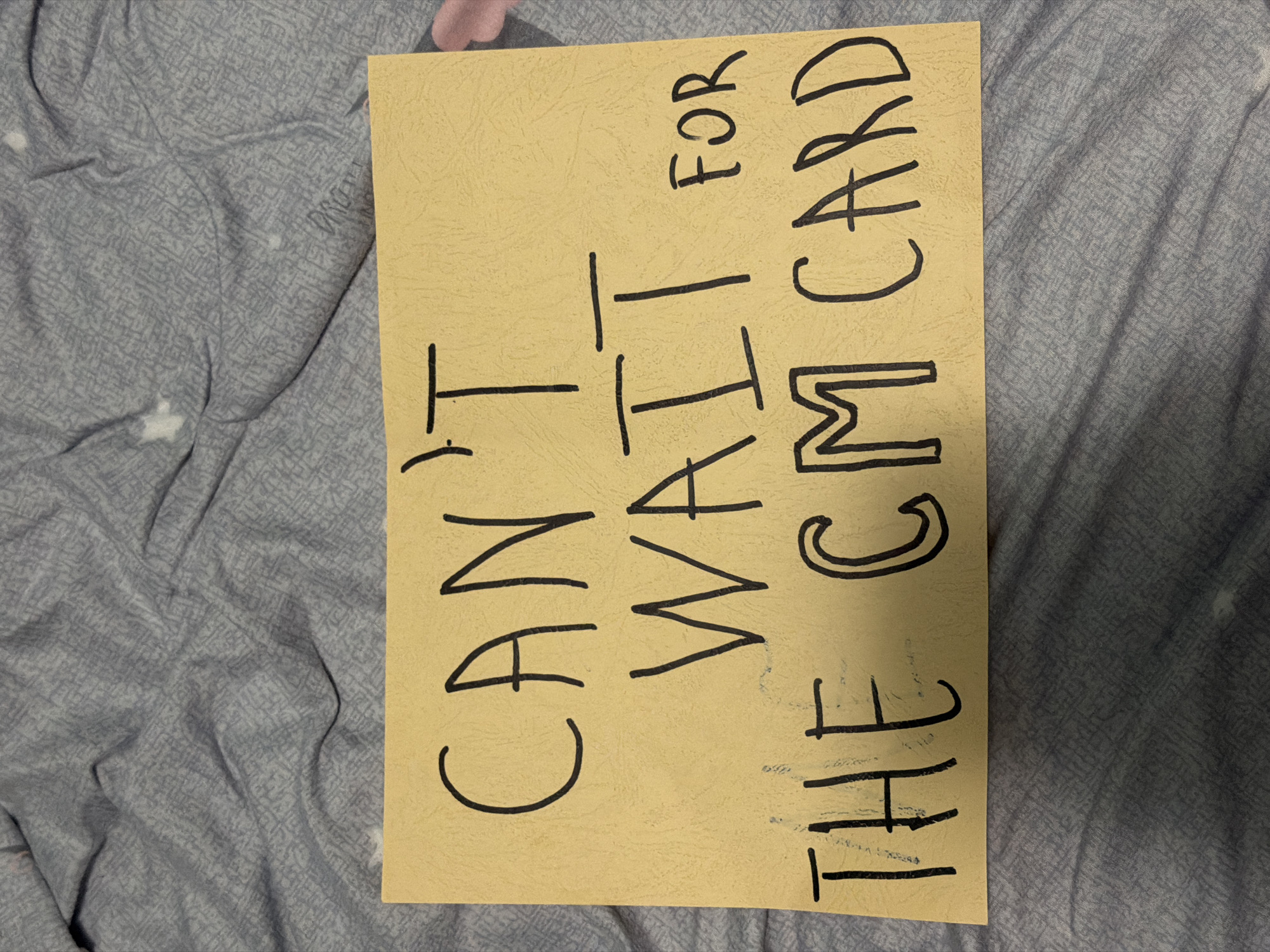 A tan colored sign with black hand-drawn lettering lies on a gray bedspread. The sign reads "Can't Wait For the CM Card". The location is unknown, although it can be said that is not Varna, Bulgaria, given the information present on the sign.