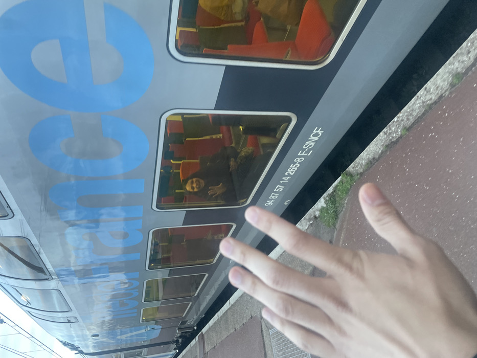 An outdoor, eye-level photograph captures a moment at a train station platform in Plaisir, France. In the foreground, the right hand of an unseen individual is extended towards a stationary double-decker train, palm open with fingers slightly spread. The train, a gray "E-SNCF" model with large blue "France" lettering along its side, is visible from its mid-section to its roofline. Through one of the train's lower windows, a female passenger with dark hair and a dark jacket is visible, seated on a red-upholstered chair, smiling and waving her right hand towards the photographer. Other red seats are visible within the train's brightly lit interior. Below the windows, the train bears the identification number "94 87 57 14 265-8 E-SNCF". The platform, composed of reddish-brown paving stones with a concrete edge, runs along the left side of the train. A metal grate is embedded in the platform to the left. A narrow strip of green grass is visible between the platform's edge and the train's lower body. The sky, appearing overcast, is reflected in the train's windows and upper metallic surface.
