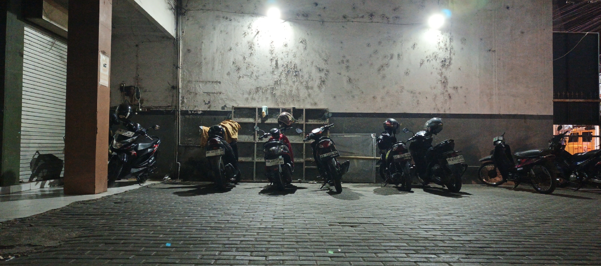 A line of parked motorcycles is illuminated by overhead lights in a dimly lit urban setting. The motorcycles are parked on a patterned brick walkway in front of a weathered, textured wall. Some of the motorcycles have helmets placed on them. To the left of the motorcycles, a large roll-up door is visible. The scene appears to be an outdoor area in Godean, Indonesia, possibly a parking lot or alleyway.