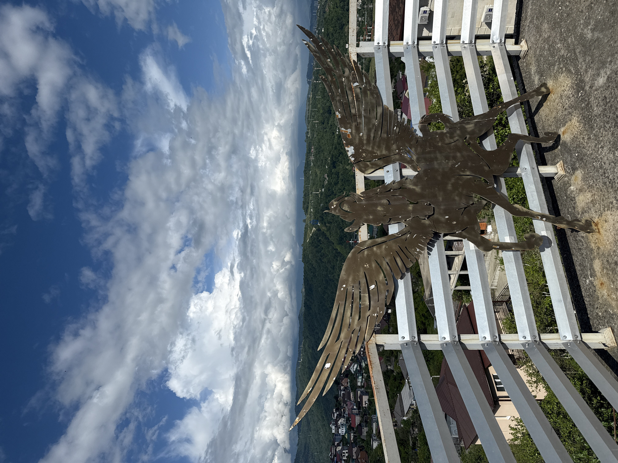 A low-angle, rotated, long-distance shot presents a bronze-colored metal statue of Pegasus mounted on a grey metal railing against a scenic backdrop. The location is likely Sochi, Russia, as evidenced by the mountain range and city visible in the background.
The statue, exhibiting a degree of weathering and rust, is positioned on the railing. The railing itself is comprised of square metal posts and horizontal slats.
The backdrop showcases a valley filled with dense vegetation, along with scattered buildings and houses. Distant mountains are veiled in atmospheric haze, while a bright sky filled with cumulus clouds dominates the upper portion of the frame.