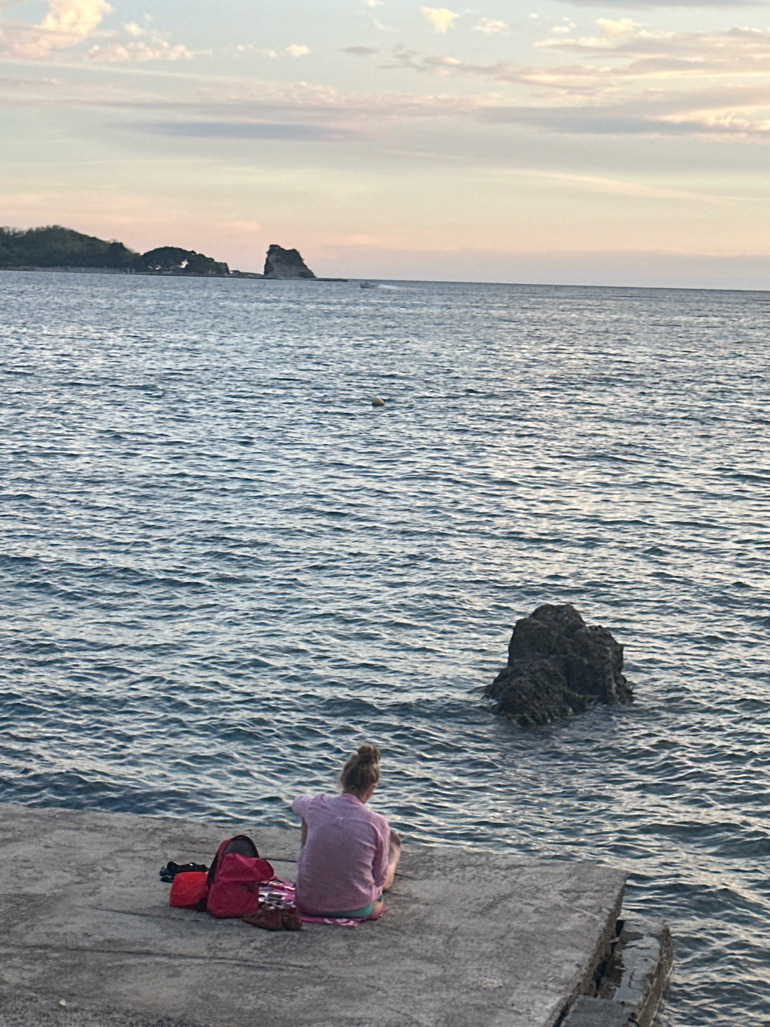 A person sits on a concrete dock overlooking the Adriatic Sea at sunset. The person is viewed from behind, wearing a light purple shirt and blue shorts. A red backpack and a striped cloth are on the dock beside them. In the water, a large dark rock is visible to the right of the person. In the distance, islands with vegetation and rock formations are visible. The sky is a soft gradient of pink, orange, and blue with wispy clouds. The scene appears to be near Budva, Montenegro, likely on the promenade or a similar waterfront area.