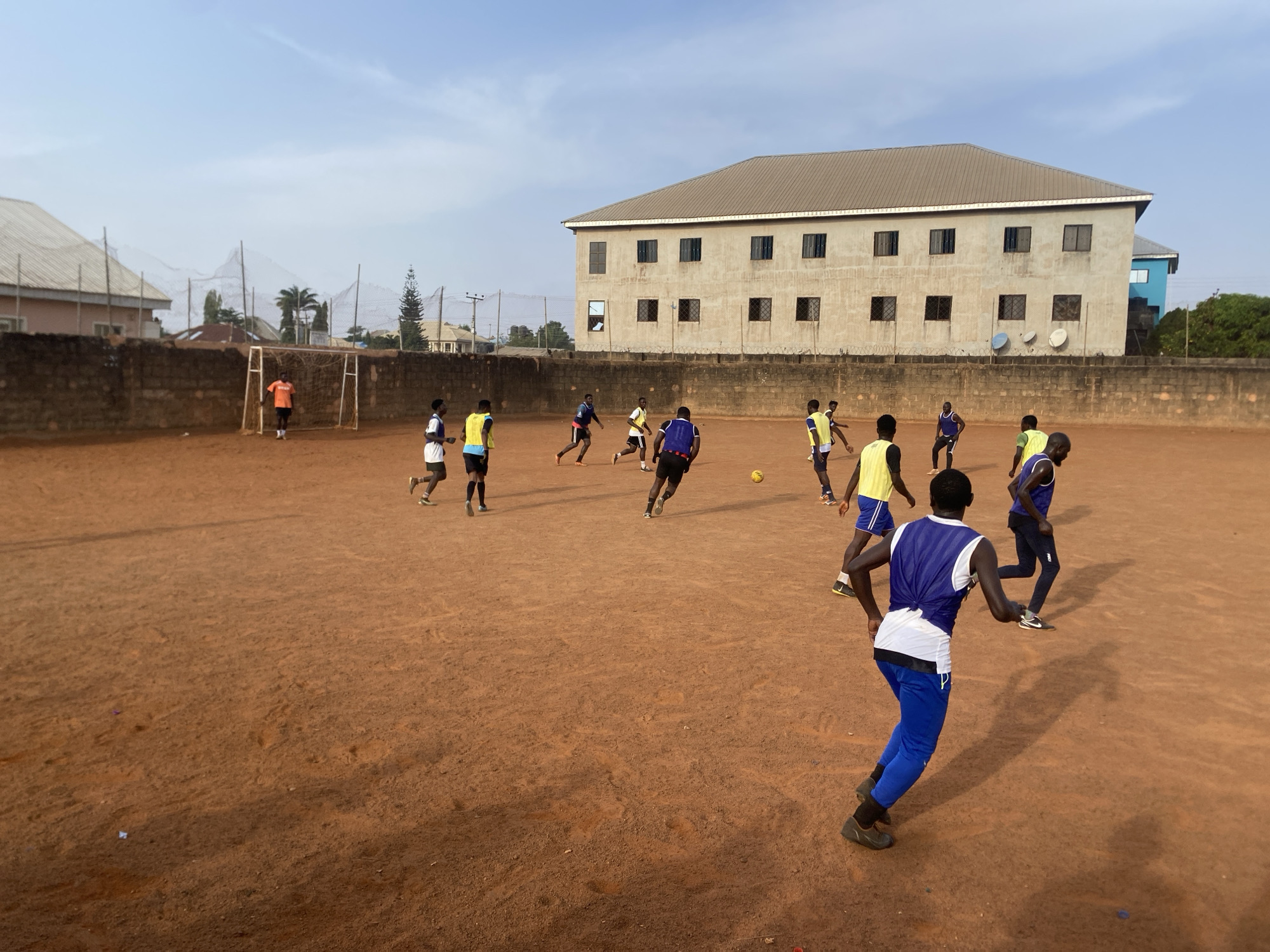 The image depicts a lively soccer game or practice taking place on a reddish-brown dirt field in Barakin Lerawi, Nigeria. Approximately 15 to 20 young men and youths are actively engaged in play, distributed across the unpaved ground. Many wear athletic gear, including colored vests or bibs in shades of yellow, blue, and orange over their shirts, along with shorts or track pants. A yellow soccer ball is visible in the mid-ground, indicating the ongoing action.
On the left side of the field, a white-framed soccer goal stands, with a player in an orange shirt positioned near it, likely serving as a goalkeeper. The players cast long shadows across the dusty surface, suggesting the time of day is either early morning or late afternoon under a clear, bright sky with minimal clouds.
The background reveals an urban setting. To the far left, residential buildings with light-colored roofs and some scattered trees are visible behind a partial wall. The dominant feature in the background is a large, multi-story concrete building with a grey roof and numerous windows, some of which are boarded or grated. This building is separated from the field by a tall brick wall topped with barbed wire, and several satellite dishes are mounted on or near it. A glimpse of another building with a blue facade is visible behind the larger grey structure.
The scene conveys an energetic community activity. Brand names "Puma" are visible on one player's tracksuit bottoms, and "NIKE" on another's shoes.