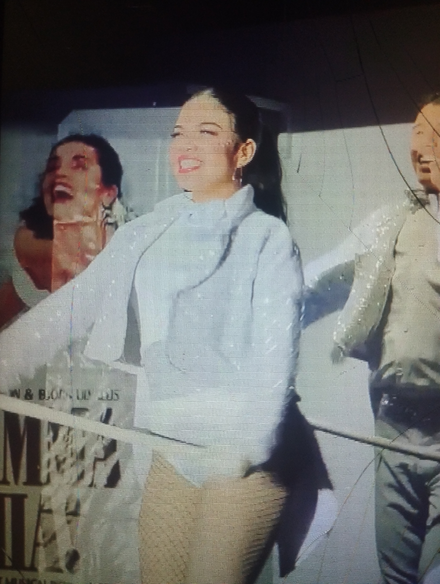 The image appears to be a photograph of a screen displaying a scene from the musical "Mamma Mia!". Three people are visible: a woman on the left laughing with her mouth open, a woman in the center wearing a white outfit with a jacket, and a man on the right also laughing with his mouth open. The background features part of the "Mamma Mia!" promotional material, including lettering. The image is grainy and shows signs of being taken of a screen, possibly a television or computer monitor. There is also some apparent damage or distortion in the image, such as scratches. The location of Punta de Piedras, Venezuela is not evident from the visual information provided in the image.