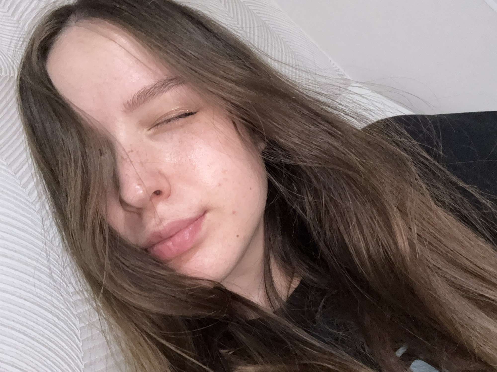 The image captures a close-up, angled portrait of a young woman resting indoors. She has long, light brown hair, some strands falling across her face. Her right eye (from her perspective) is closed, while her left eye is mostly obscured by her hair. A gentle, serene smile plays on her naturally glossy, pink lips.
Her fair skin exhibits a natural complexion with a few small freckles or moles visible on her cheek and chin. She appears to be wearing a dark, possibly black, top, a portion of which is visible. She is positioned against a textured, light-colored surface, likely a pillow or bedding, on the left side of the frame, and a dark, indistinct background on the right. The lighting is soft and diffused, highlighting a slight sheen on her skin and lips, suggesting a relaxed or comfortable environment. The overall impression is one of peace and tranquility. There is no visible text in the image. No specific time of day or overt location cues are present, beyond the general indoor setting. The location context provided is Chaikovsky, Russia.