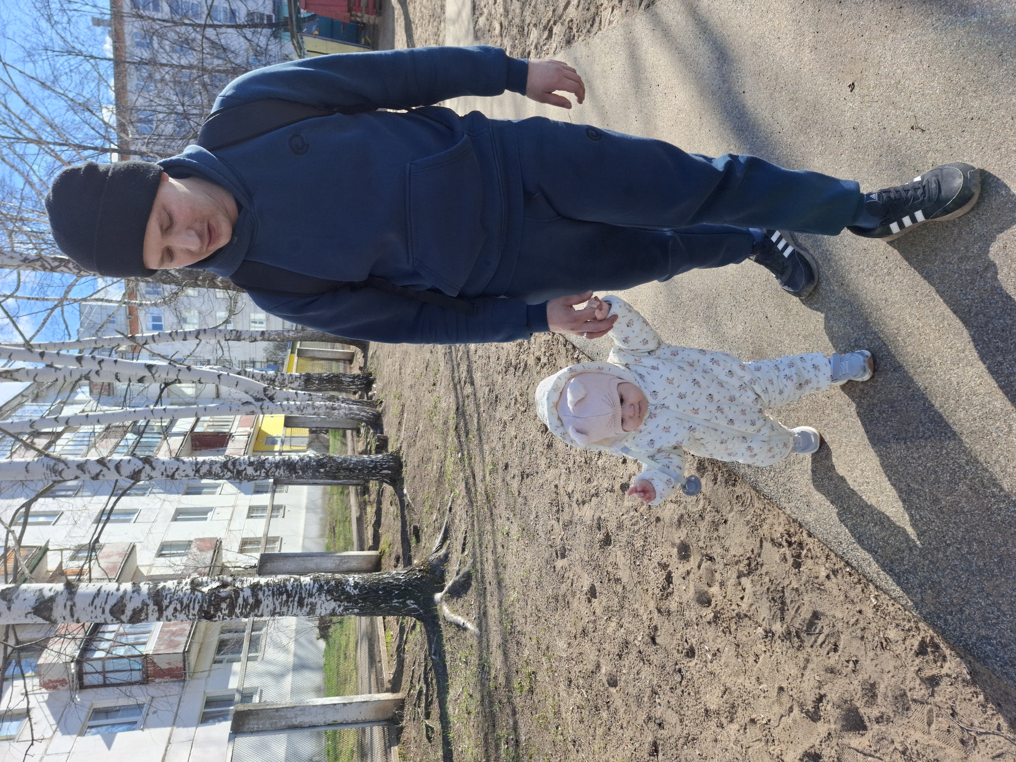 A man and a baby are walking on a sidewalk in Chaikovsky, Russia. The man is wearing a black beanie, a dark blue hoodie and sweatpants, and black Adidas sneakers with white stripes. He also has a black backpack. The baby is wearing a light-colored patterned onesie and a light pink hat. They are holding hands. To the left of them are birch trees and a residential building. The ground is a mix of sidewalk and dirt. The picture appears to be taken during the day with sunlight present.