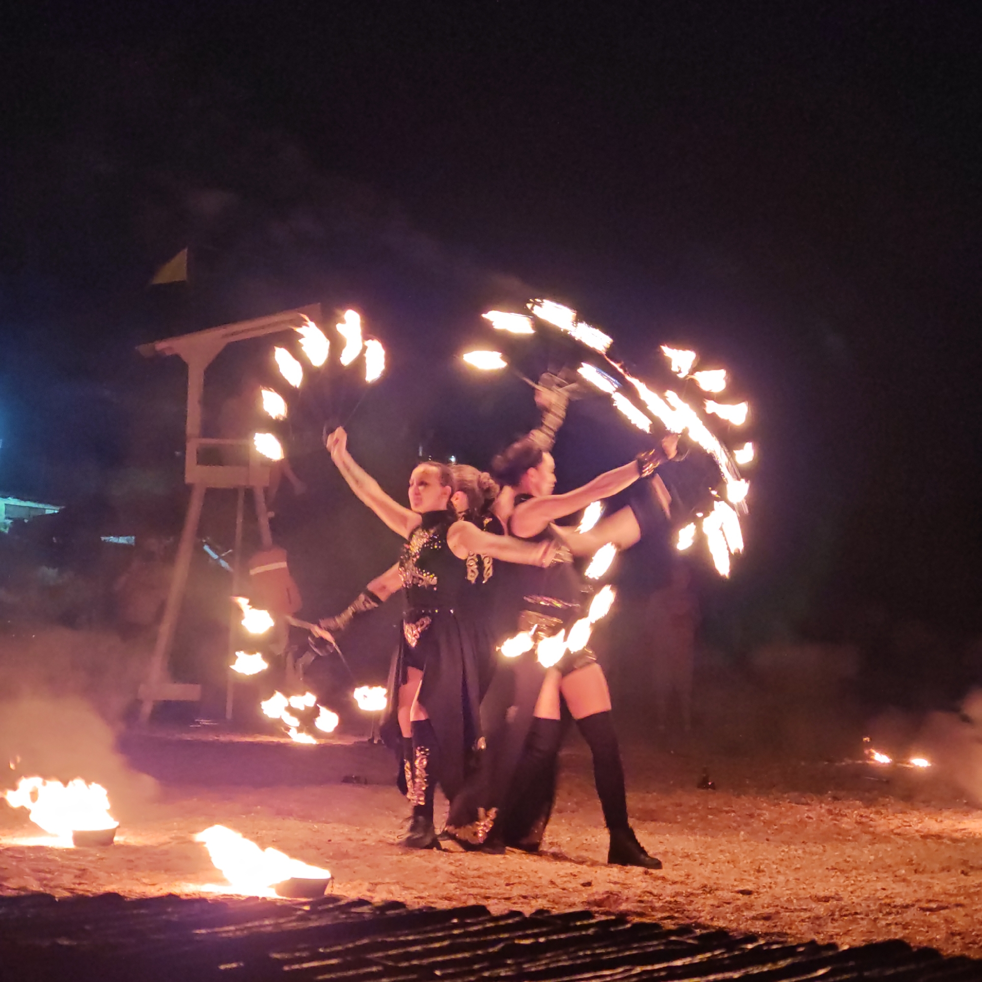 The image depicts a night scene in what appears to be an outdoor performance area in Shchyolkino, Ukraine. The primary focus is on three women engaged in fire dancing. They are dressed in dark, elaborate costumes with embellishments and are holding fire props, creating circular patterns of flames around them. The ground appears to be covered with sand or dirt, with several buckets of fire also visible, contributing to the illumination of the scene. In the background, a wooden structure, possibly part of a stage or prop, stands with flames emanating from it. The overall atmosphere is one of a lively evening performance, with the darkness of the night contrasted by the bright, dynamic flames.