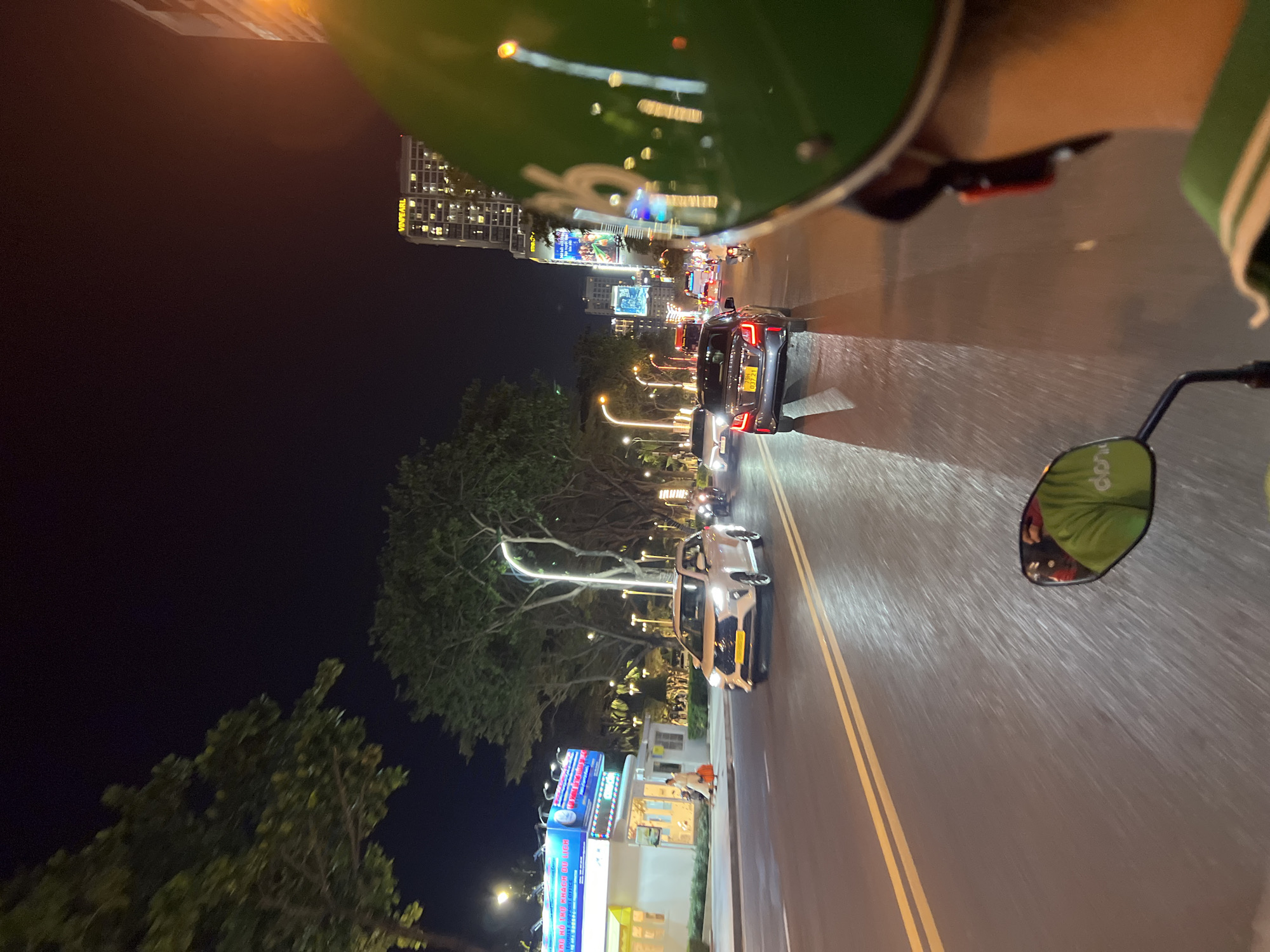 The photograph captures a nighttime scene in Nha Trang, Vietnam, from the perspective of someone riding a scooter. The scooter is traveling on a well-lit road with double yellow lines. A dark sky contrasts with the city lights in the distance. A row of trees lines the left side of the road, while buildings and streetlights illuminate the right side. Traffic consists of several cars, including a gray car with a license plate visible in the picture, moving in the same direction. The reflection of the streetscape can be seen in the shiny helmet and side mirror of the scooter. A building with illuminated signs can be seen on the roadside.