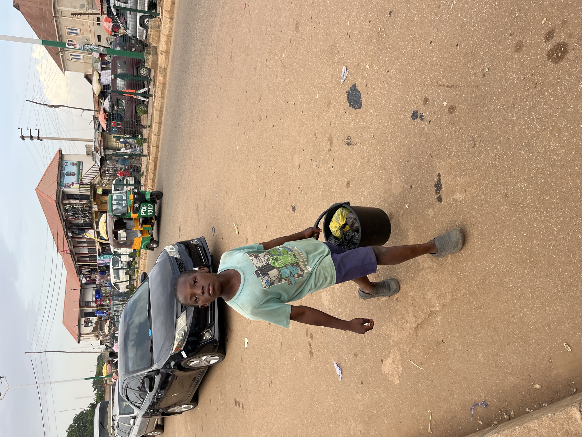 In Bukuru, Nigeria, a young boy walks on a dusty street, carrying a black bucket filled with what appears to be trash. He is wearing a light blue t-shirt with a graphic print and dark blue shorts, along with gray shoes. The street is lined with shops and market stalls, selling various goods under awnings. Several vehicles are present, including a black sedan, a white minivan, and a yellow and green auto-rickshaw. The sky is overcast, suggesting a cloudy day. The overall scene depicts a busy, commercial area in an urban environment.