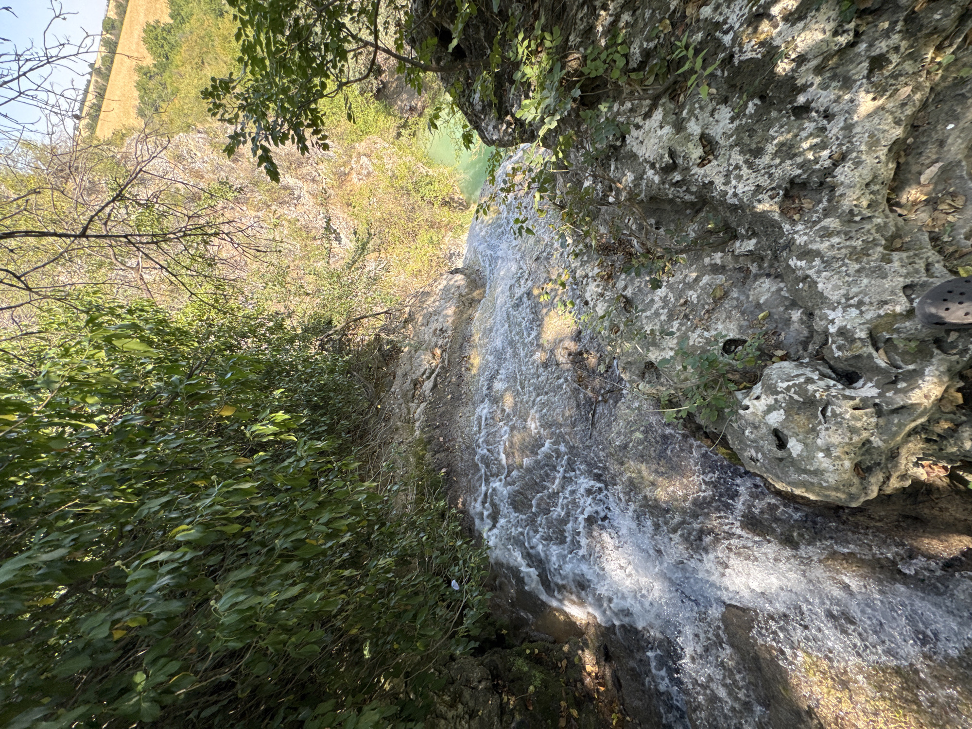 The image shows a top-down view of a waterfall cascading over rocks in the Hotnitsa Waterfall area, Bulgaria. Lush green foliage lines the left side of the waterfall, with branches extending into the frame from the top. The water is white and frothy as it flows down, with sunlight glinting off its surface. The rocks are uneven and grey, with some patches of moss and dry leaves. In the distance, beyond the waterfall and foliage, a turquoise pool of water is visible, surrounded by dry, sun-drenched hills and scattered green trees. The overall scene depicts a natural, outdoor environment. A dark, textured shoe is visible at the bottom right corner of the frame, suggesting a person is present at the edge of the waterfall.