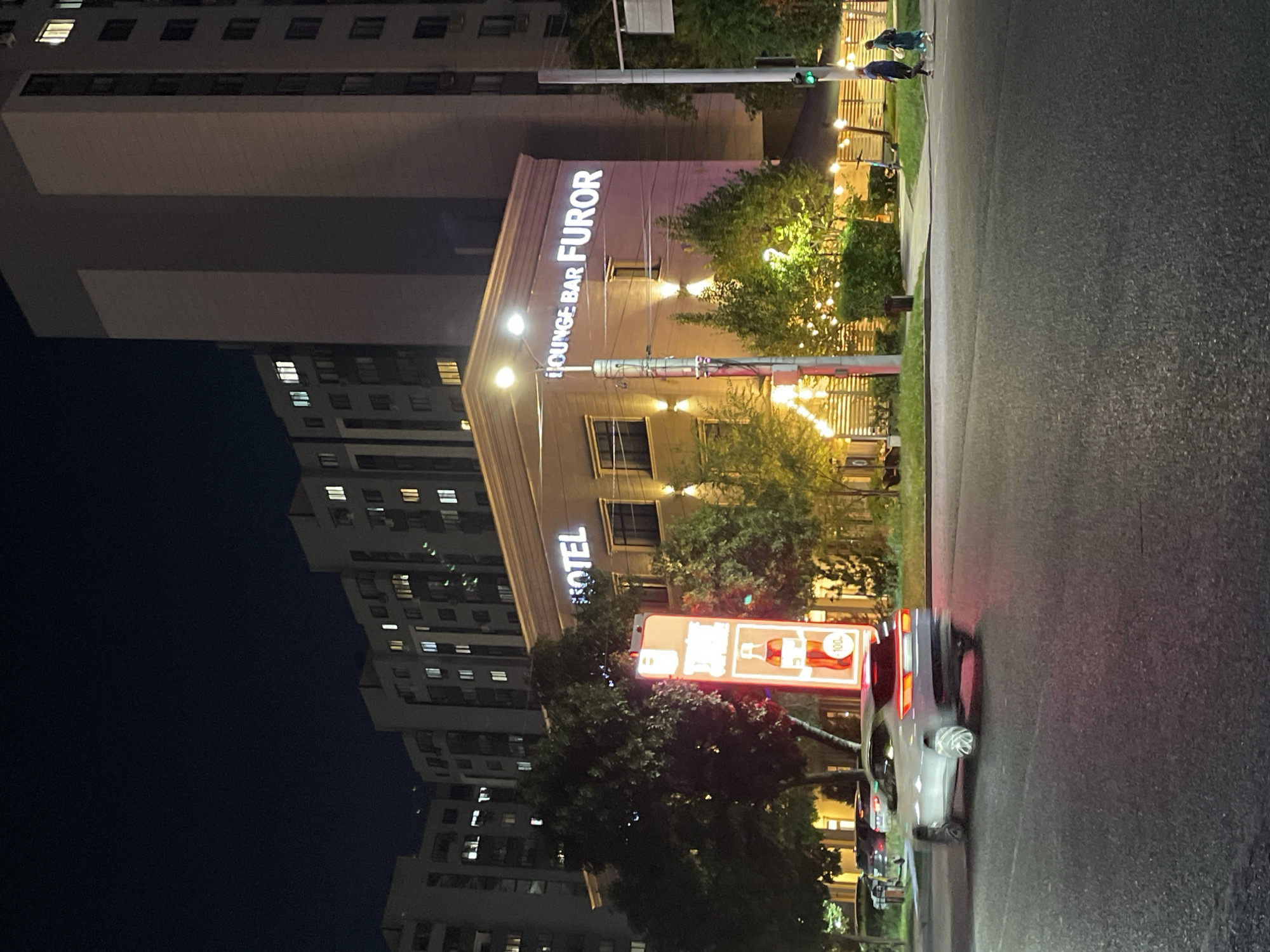 This is a night-time, medium-shot photograph taken from a street intersection in Almaty, Kazakhstan. In the foreground, a light-colored car is captured in motion, appearing to turn. The road surface is asphalt. Behind the car, a brightly lit vertical advertisement sign is visible. To the right of the sign, a multi-story building is illuminated, with a prominent sign reading "HOTEL". Further to the right, another building features a sign that reads "LOUNGE BAR FUROR". Trees with foliage are present in front of these buildings, and some are adorned with small lights. In the background, several tall apartment buildings are visible, with many windows illuminated. The sky is dark. Two individuals are walking on the sidewalk on the far right of the image, near a traffic light which is illuminated green.