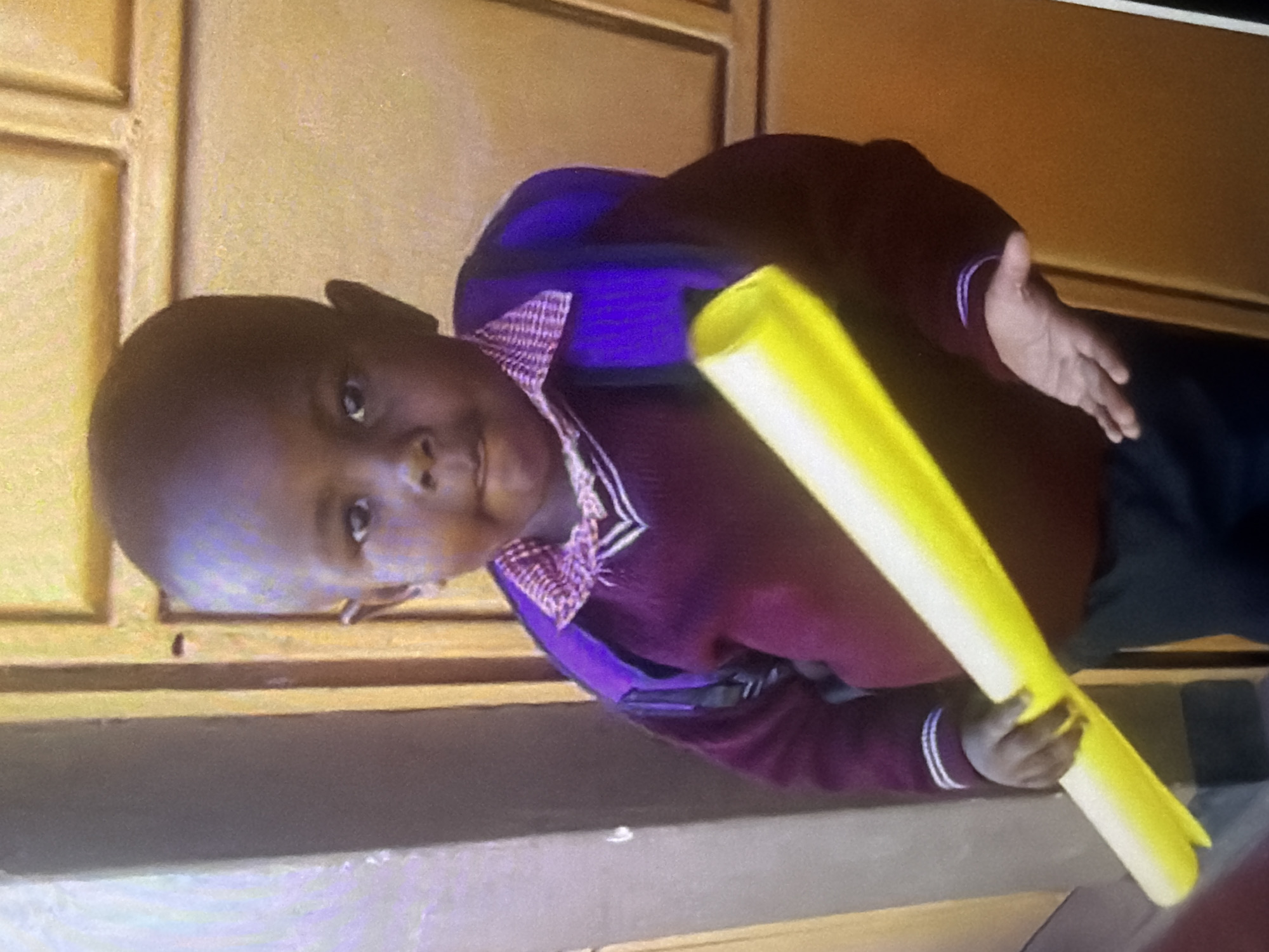 In an indoor setting with a yellowish-brown door in the background, a young child of African descent, with dark skin and short, dark hair, stands facing forward. The child wears a burgundy sweater with a purple collar and trim, and a backpack that is partially visible and appears to be blue. In their hands, the child holds a yellow cylindrical object, possibly a pool noodle or a poster tube. The child's left hand grips the object, while their right hand is open and extended downwards. The lighting is somewhat dim, with a slight glare on the subject.