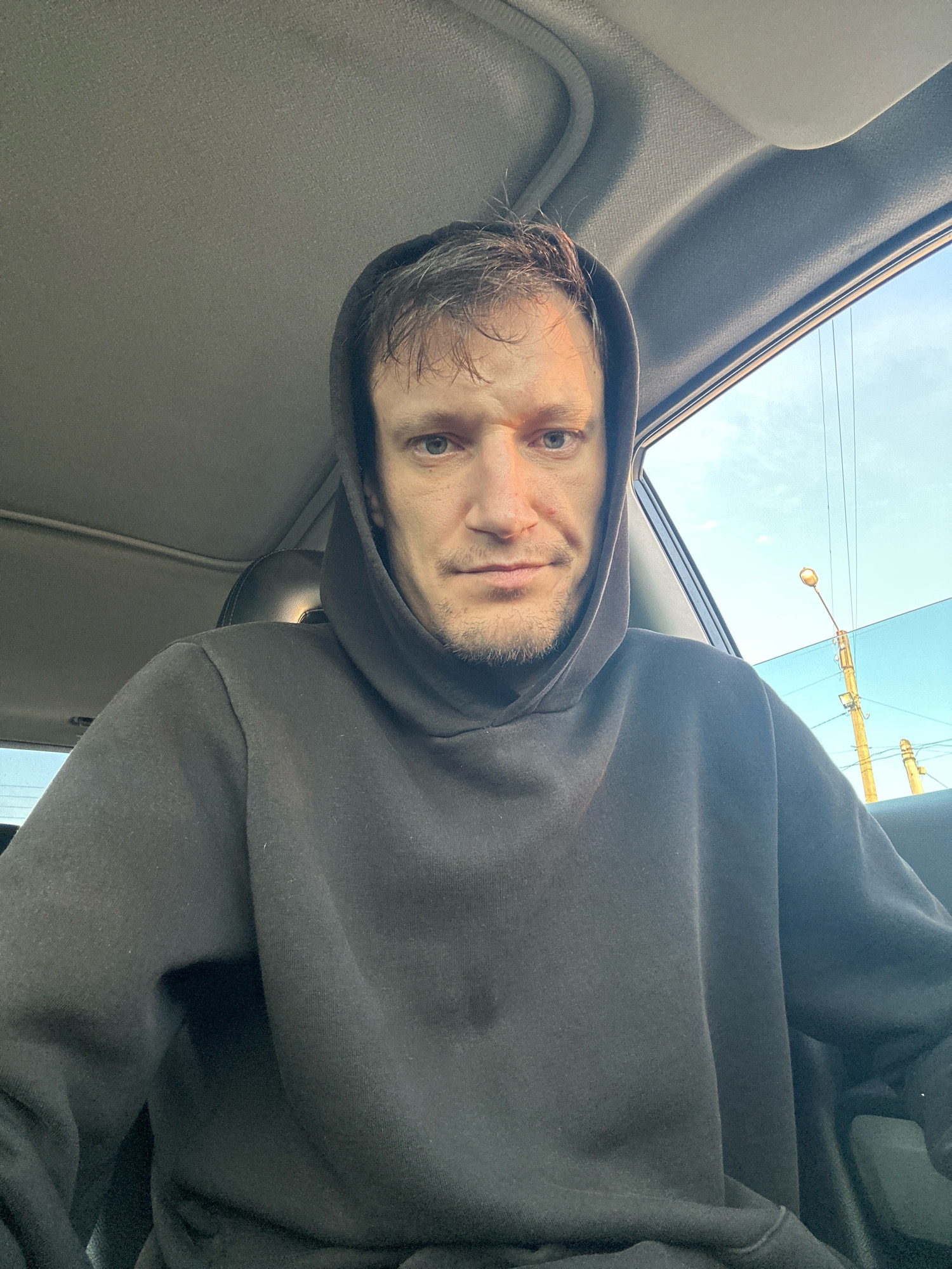 A medium shot selfie taken from inside a vehicle shows a Caucasian male with short, messy brown hair and light blue eyes. He is wearing a dark grey hooded sweatshirt with the hood up, obscuring the top of his head. His face is centered in the frame, looking directly at the camera with a neutral expression. He has stubble on his chin and a faint scar above his left eyebrow.
The interior of the vehicle is visible, with the grey headliner and a portion of the car's roof. Sunlight streams in from the right side, illuminating the man's face. Through the open car window on the right, a clear blue sky is visible, along with power lines and a street lamp. The exterior scene suggests an outdoor urban or suburban environment. The vehicle appears to be parked.
The location is identified as Makiv, Ukraine, based on the provided context.